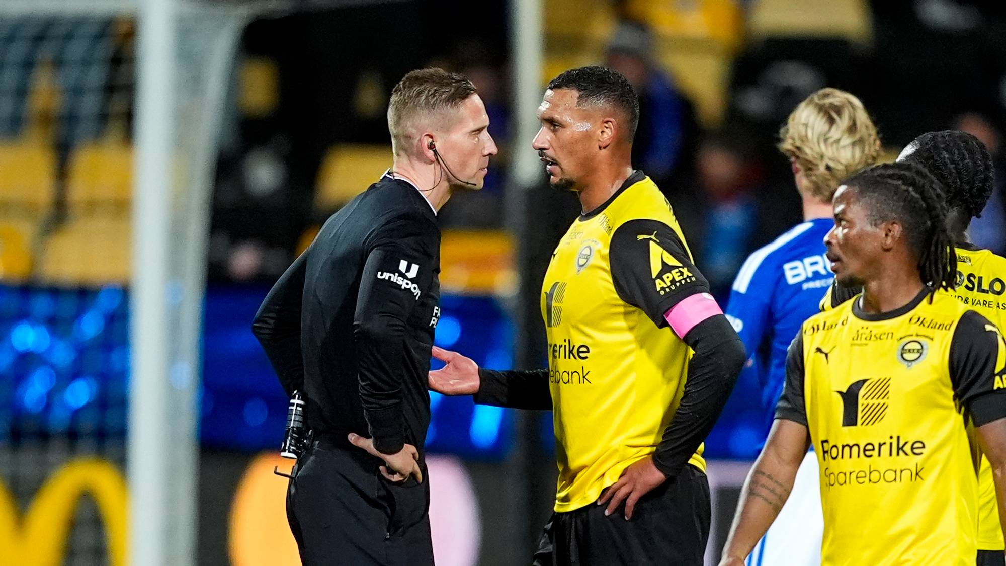 Fotballspiller diskuterer med dommer på banen mens andre spillere ser på. (Bildebeskrivelsen er laget av en KI-tjeneste)