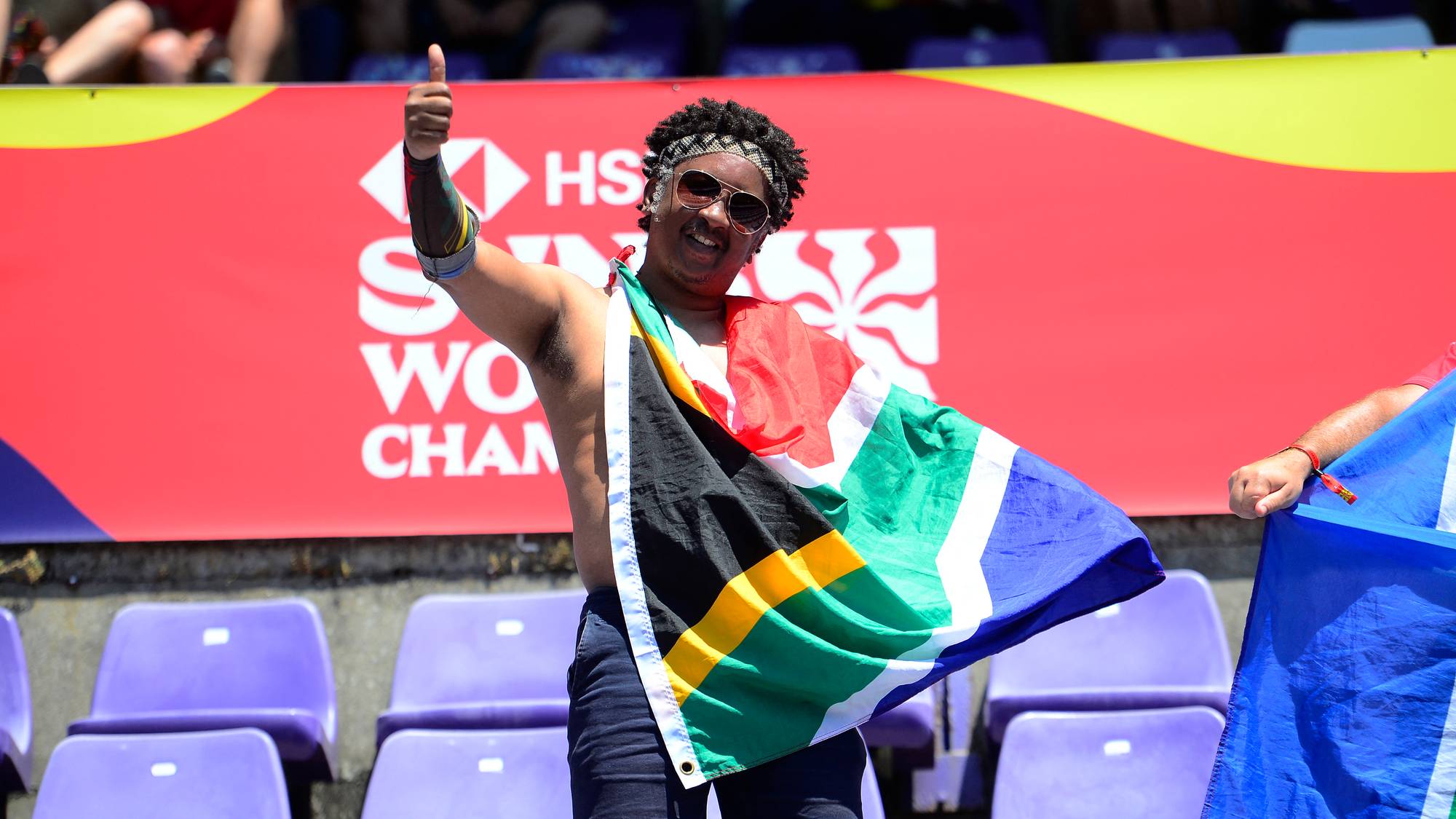 En person med Sør-Afrikas flagg gir tommel opp. Bakgrunnen viser en reklame for HSBC World Rugby Sevens Series. Personen står foran tomme tribuneseter. Et annet flagg er synlig til høyre. (Bildebeskrivelsen er laget av en KI-tjeneste)
