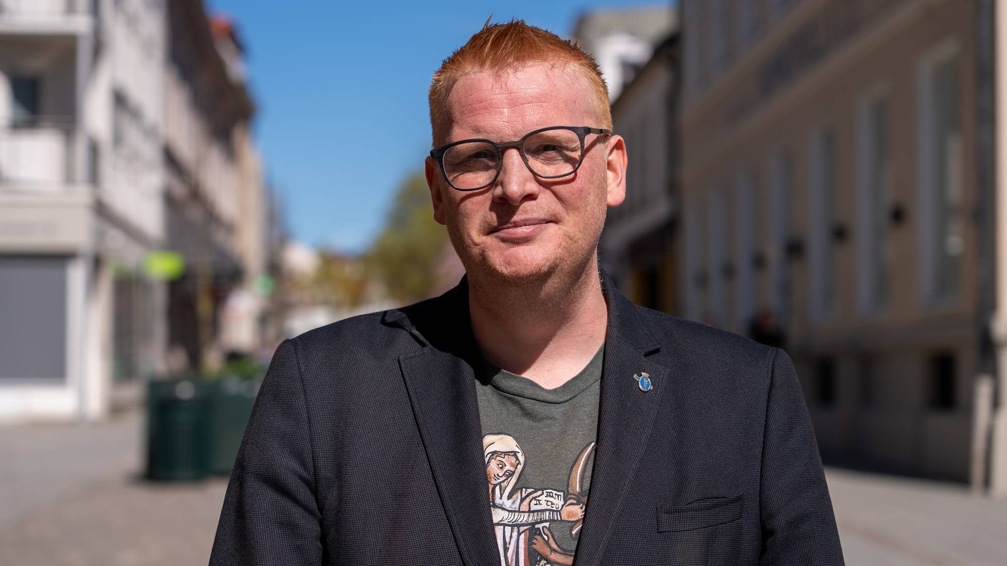 En mann med briller og rødt hår står utendørs. Han har på seg en mørk dressjakke over en t-skjorte. I bakgrunnen er en gate med bygninger på begge sider. (Bildebeskrivelsen er laget av en KI-tjeneste)