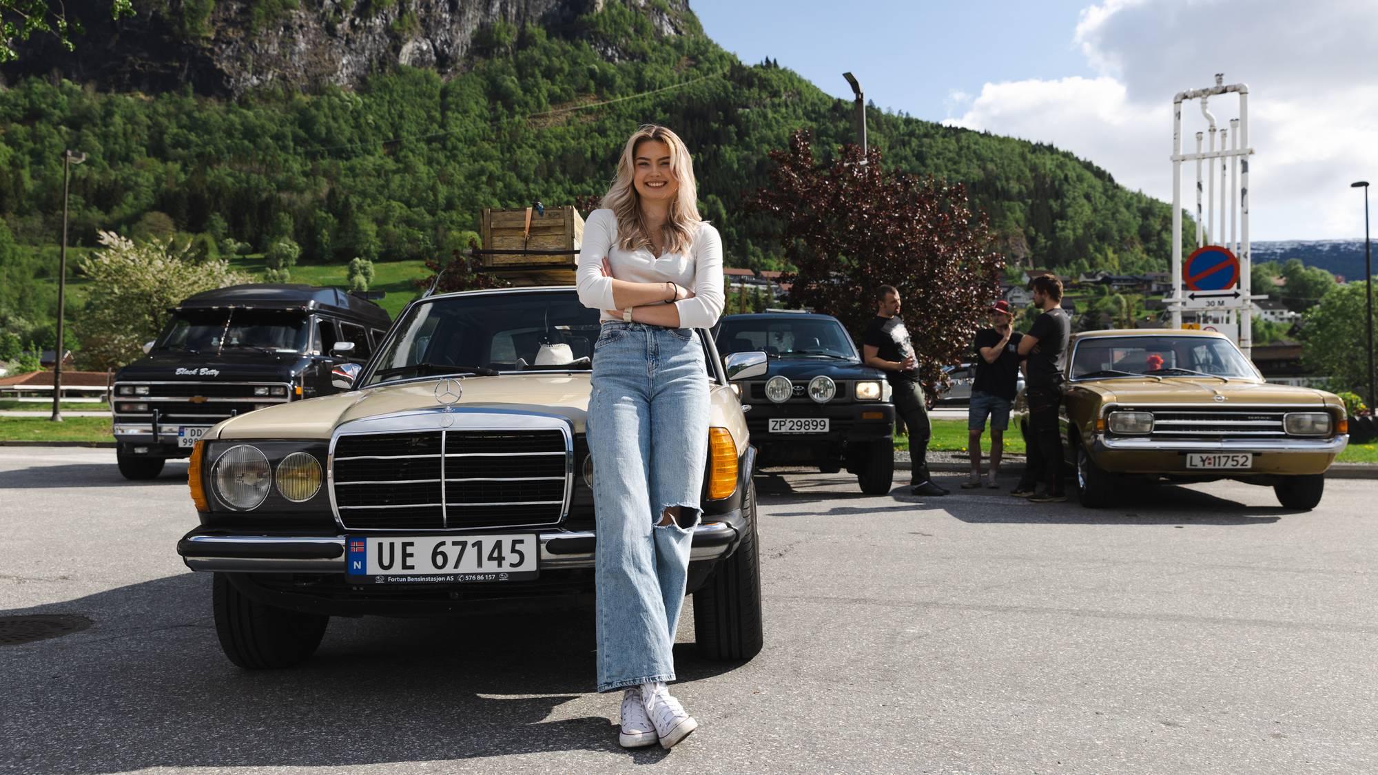 Nina sitter foran på bilen sin, en eldre Mercedes, og smiler bredt til kamera. Hun har blondt bølgete hår, en tynn hvit genser og lyseblå olabukser. Det er sol og fint vær, og vi kan se grønn skog og fjell bak Nina. Rett bak henne ser vi flere andre kule biler, både store og små. Det står en gruppe med venner og prater i bakgrunnen. 