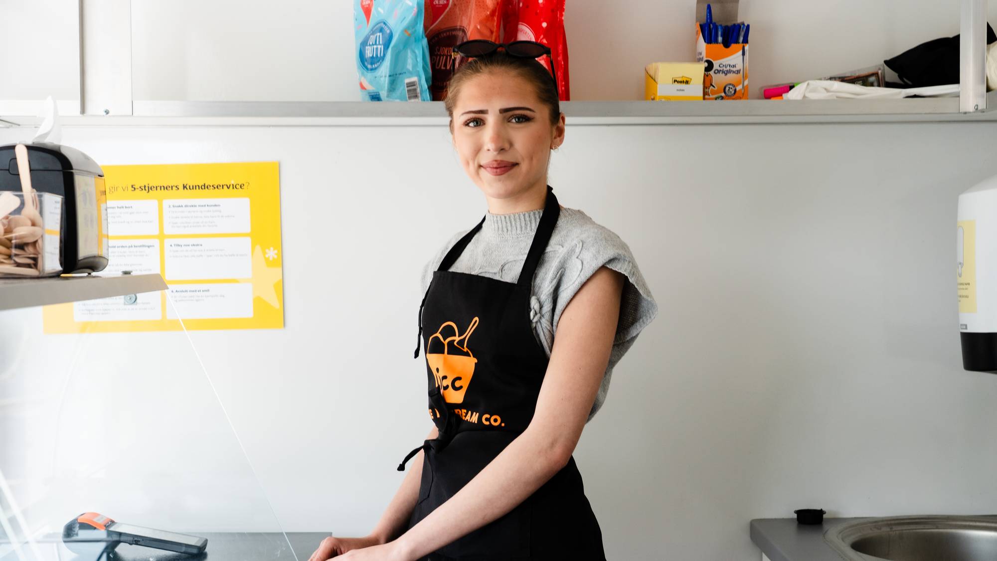 En ung kvinne i forkle står bak en disk i det som ser ut som en kiosk eller kafé. Hun smiler mot kameraet. På disken står en betalingsterminal. Bak henne er det en hylle med diverse varer. 