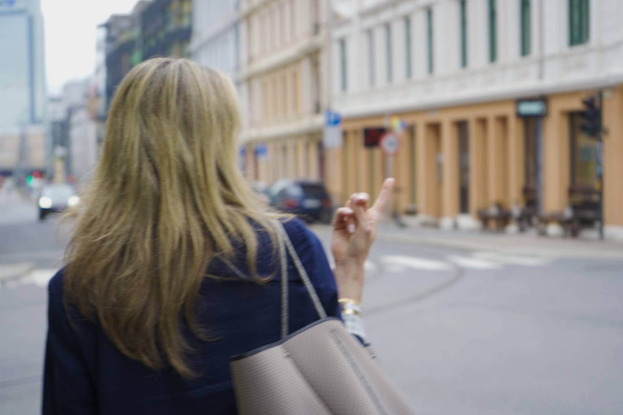 En kvinne med blondt hår holder opp en finger mens hun går over en gate. Hun bærer en mørkeblå jakke og en stor veske. I bakgrunnen er det bygninger og biler. (Bildebeskrivelsen er laget av en KI-tjeneste)