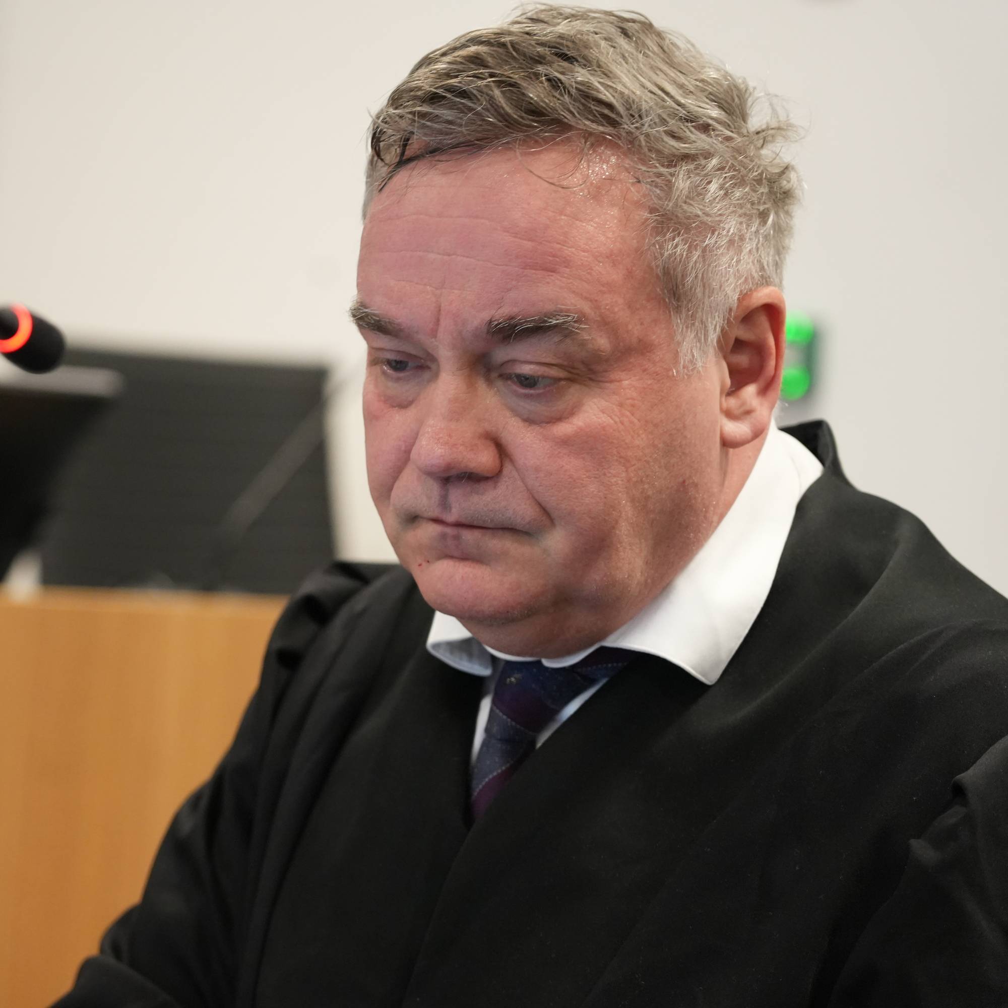 En person i dommerkappe sitter i en rettssal foran en mikrofon. Det er synlig en dataskjerm og en trebarriere i bakgrunnen. Rettsalen er nøytralt innredet med fokus på utstyr som mikrofoner og skjermer. Bildet gir inntrykk av en formell rettssituasjon. (Bildebeskrivelsen er laget av en KI-tjeneste)