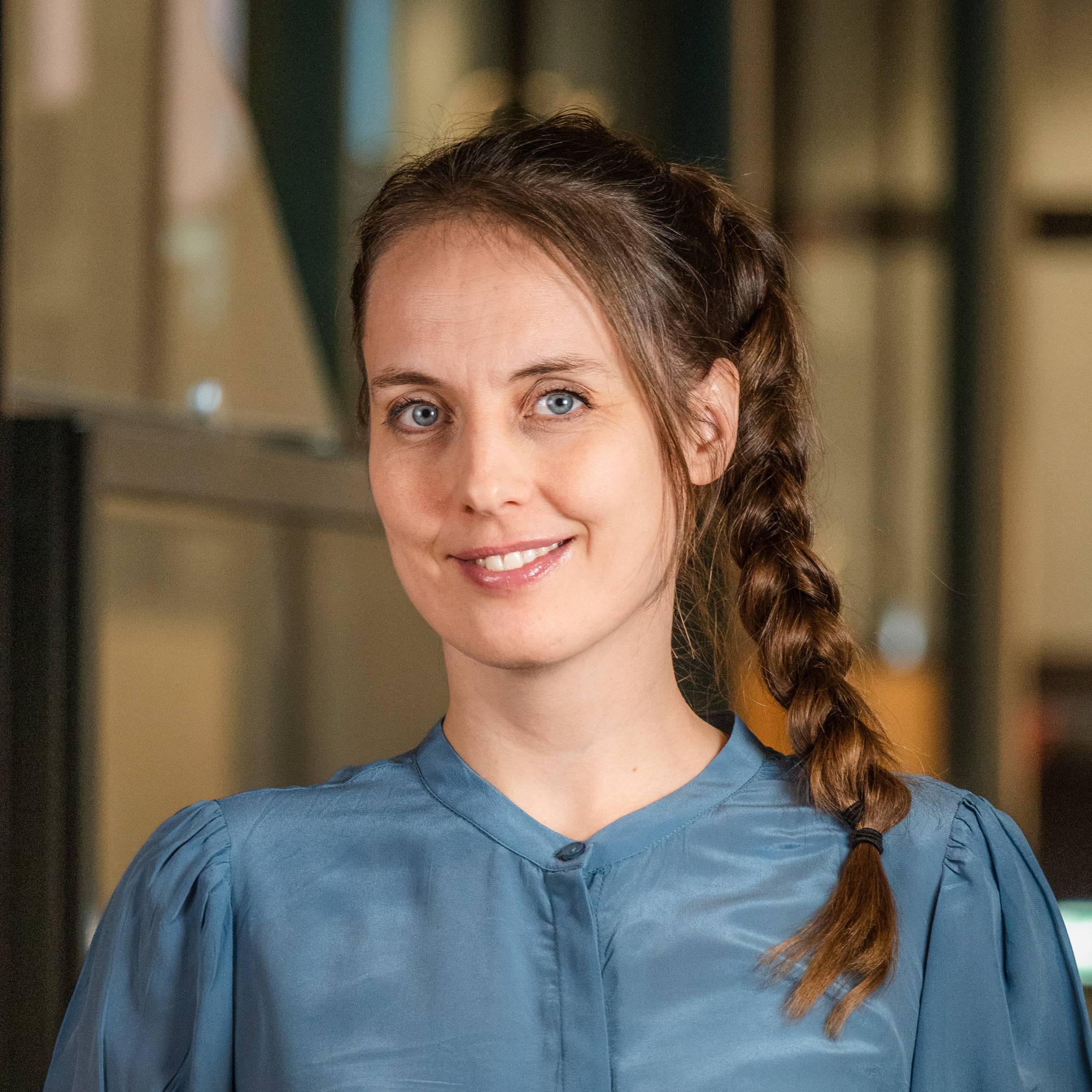 Heidi Røneid står i turkis skjorte og smiler til kameraet. Hun har brunt hår i en flette