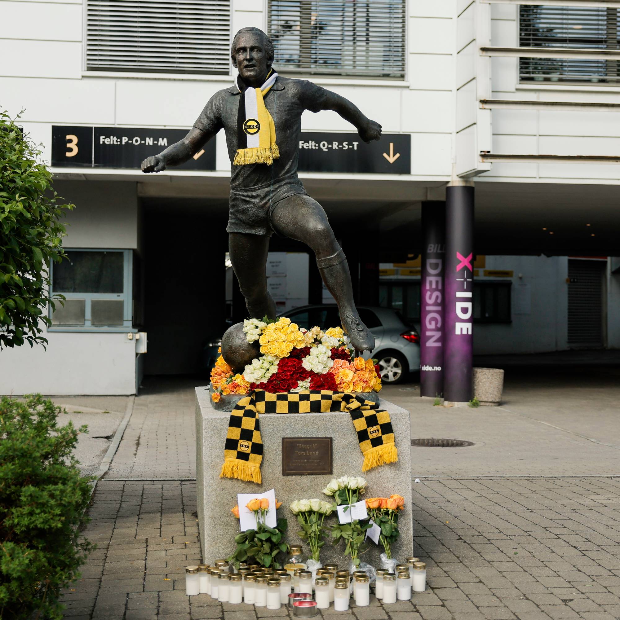 En statue av en fotballspiller står foran en bygning. Blomster og lys er plassert ved foten av statuen. (Bildebeskrivelsen er laget av en KI-tjeneste)