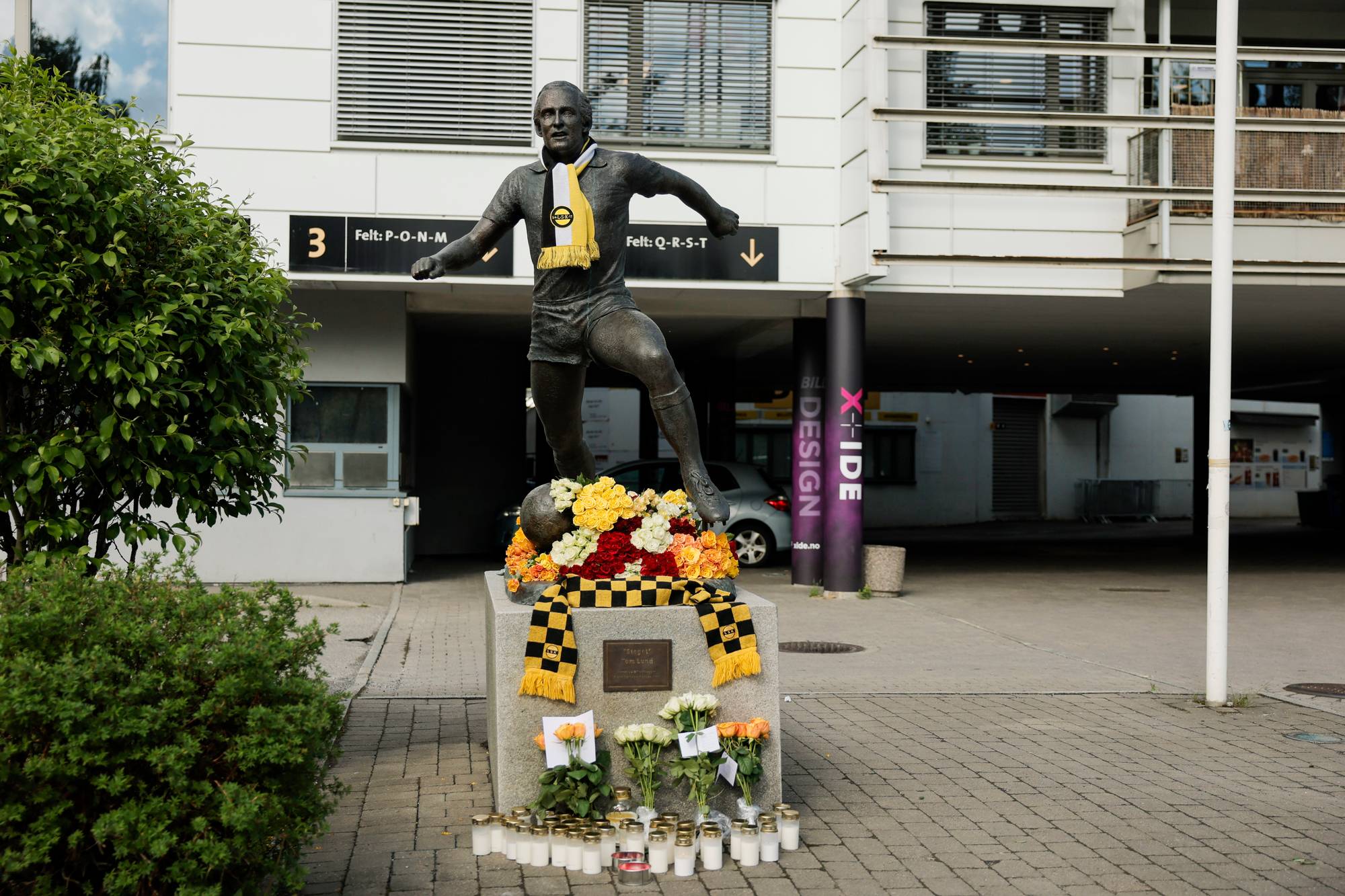 En statue av en fotballspiller står foran en bygning. Blomster og lys er plassert ved foten av statuen. (Bildebeskrivelsen er laget av en KI-tjeneste)