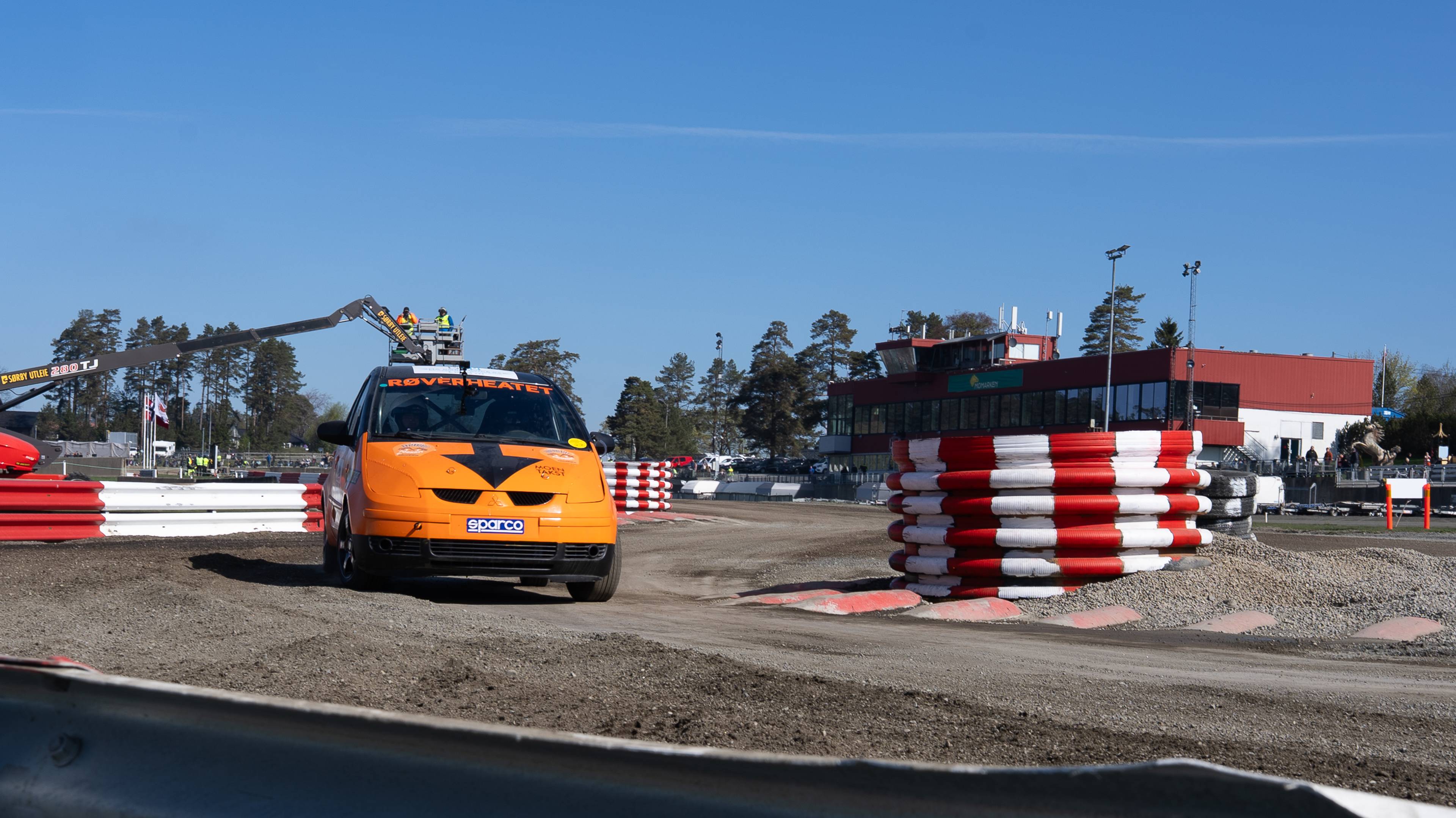 En oransje racingbil står på en grusbane. En kran med to personer i kurven står over bilen. Det er et rødt og hvitt sperringsbatteri til høyre og en bygning bak. (Bildebeskrivelsen er laget av en KI-tjeneste)