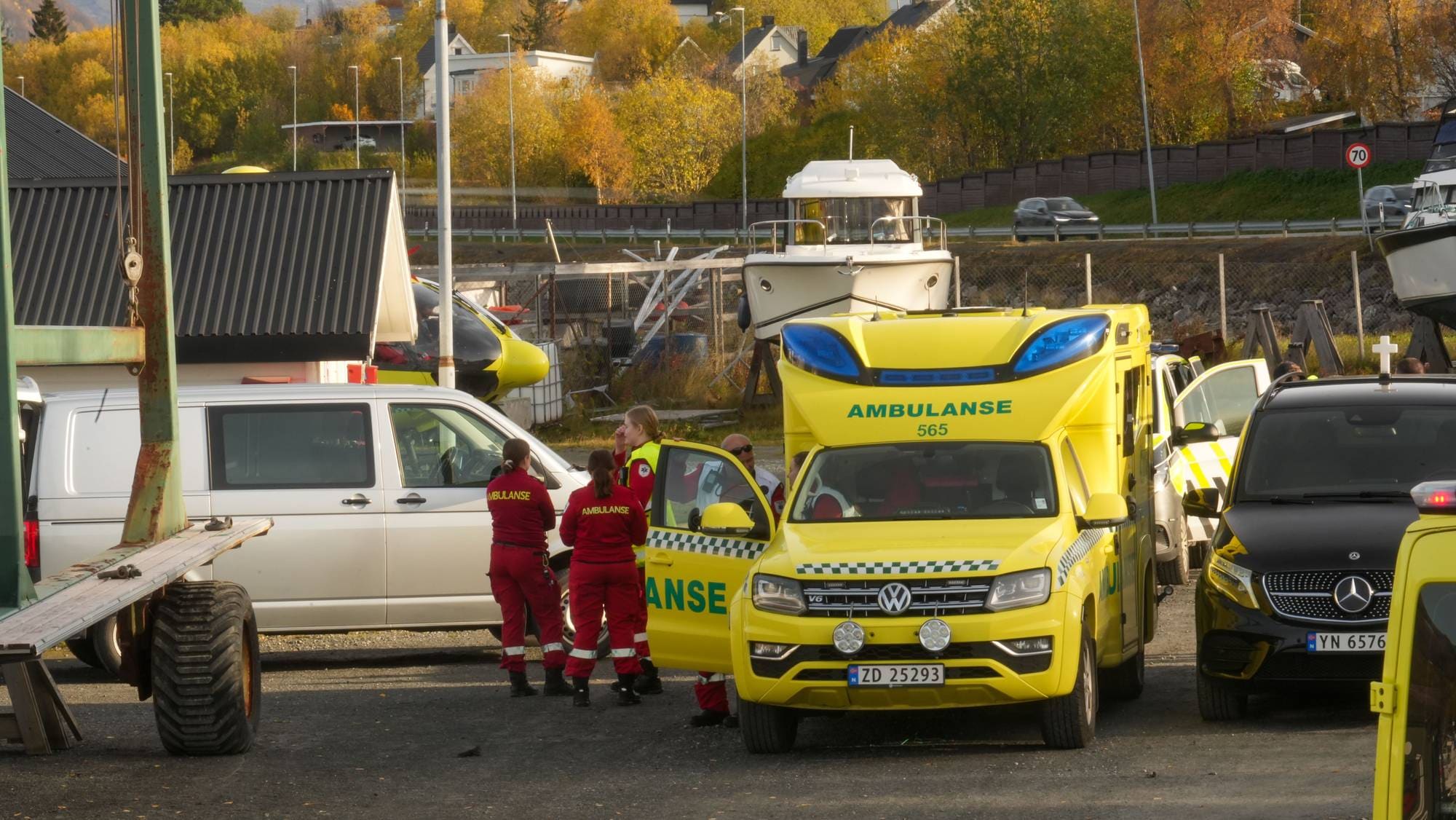 Ambulansepersonell står ved en gul ambulanse på et havneområde. Flere kjøretøy er parkert i nærheten. (Bildebeskrivelsen er laget av en KI-tjeneste)