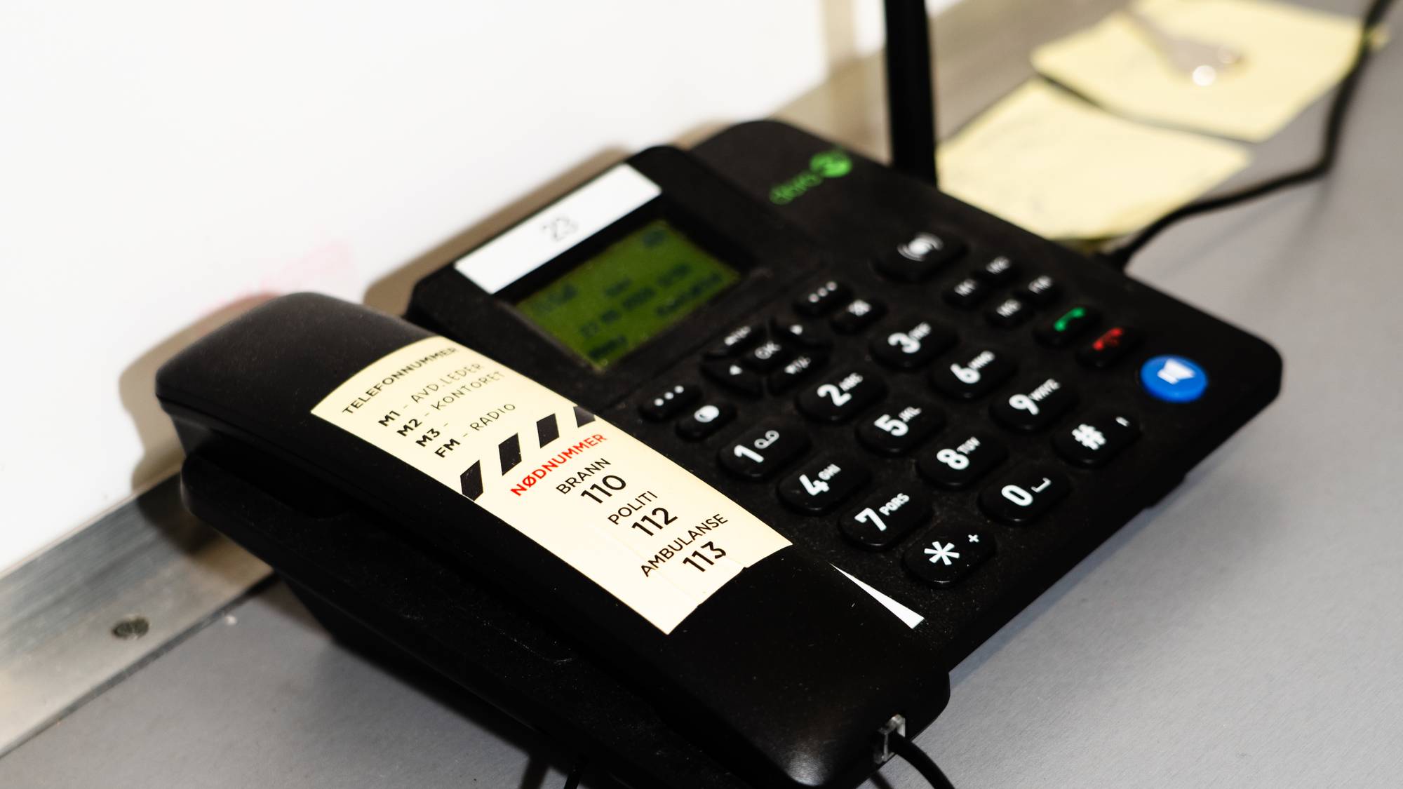 Et nødnummer-telefon står på et grått bord. Telefonen er svart, og har en ledning og en antenne. På telefonen er det en klistrelapp med nødnumre: 110 for brann, 112 for politi og 113 for ambulanse. 