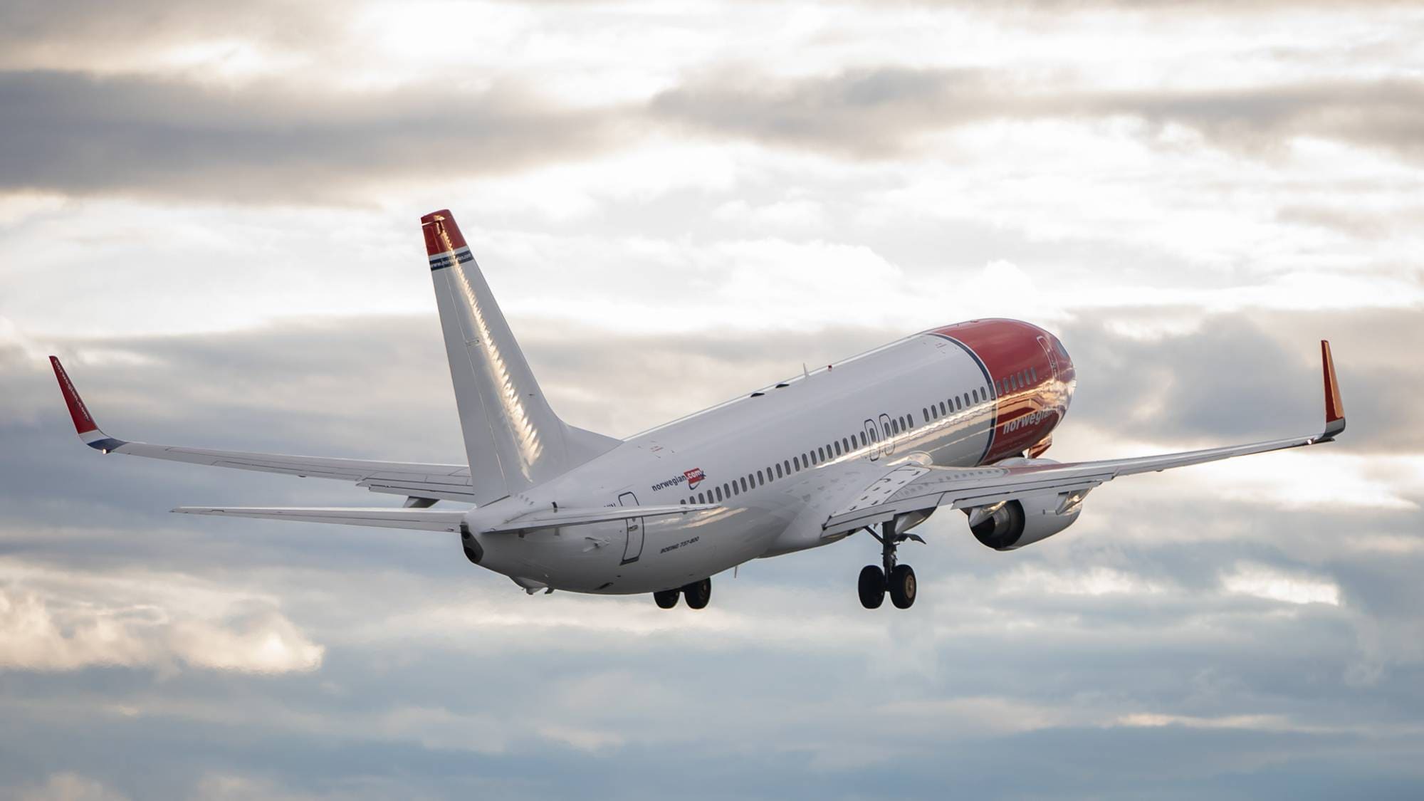 Et Norwegian-fly av typen Boeing 737-800 har akkurat tatt av fra Trondheim lufthavn Værnes. Hjulene er fortsatt ut. Himmelen med solglimt gjennom skyene omringer flyet.