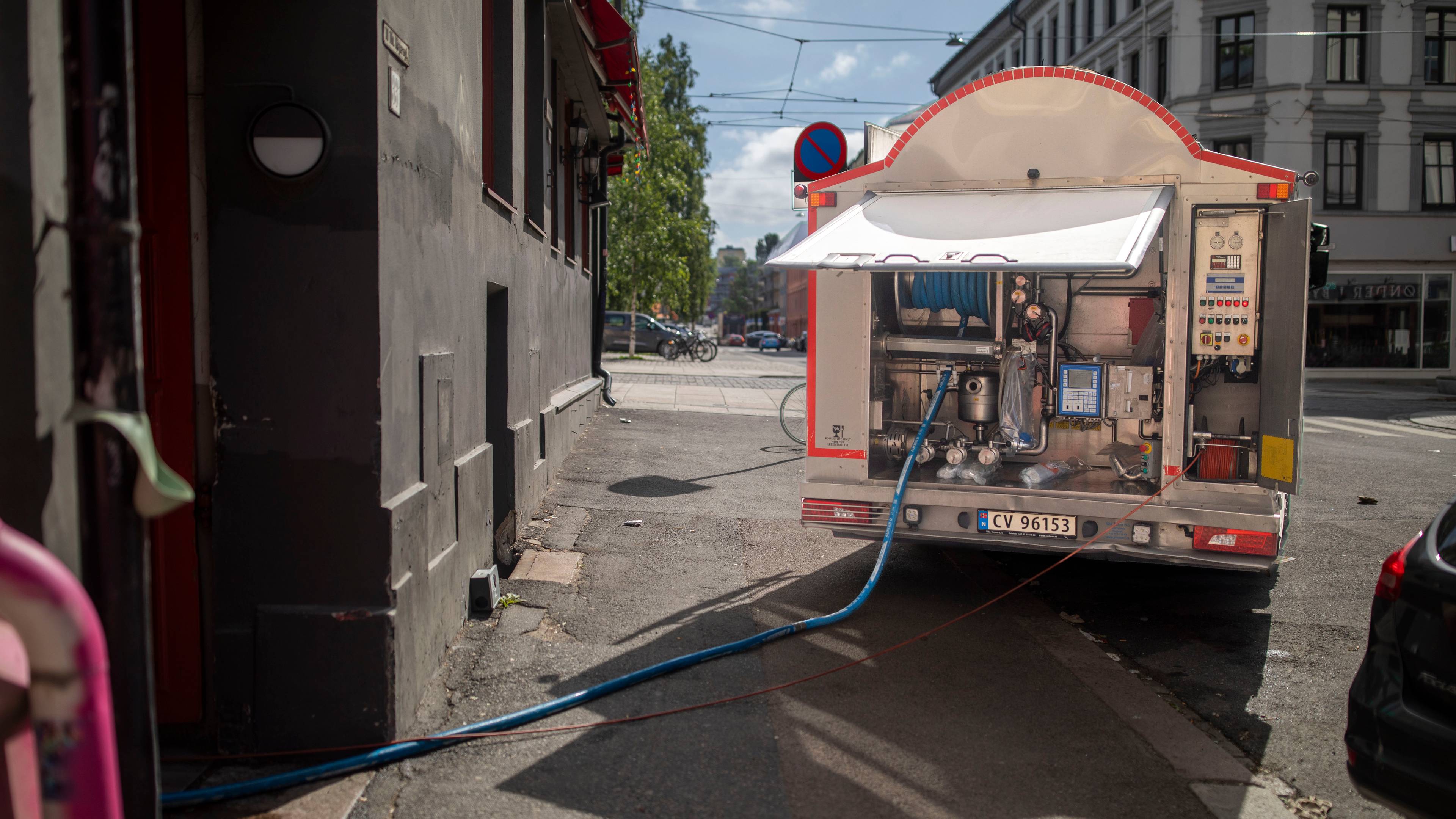 En tankbil med en blå slange koblet til bakenden er parkert ved en bygning. Bilen er åpen og viser utstyr for væskehåndtering. En annen slange ligger langs bakken. Bakgrunnen viser et bybilde med bygninger og trær.