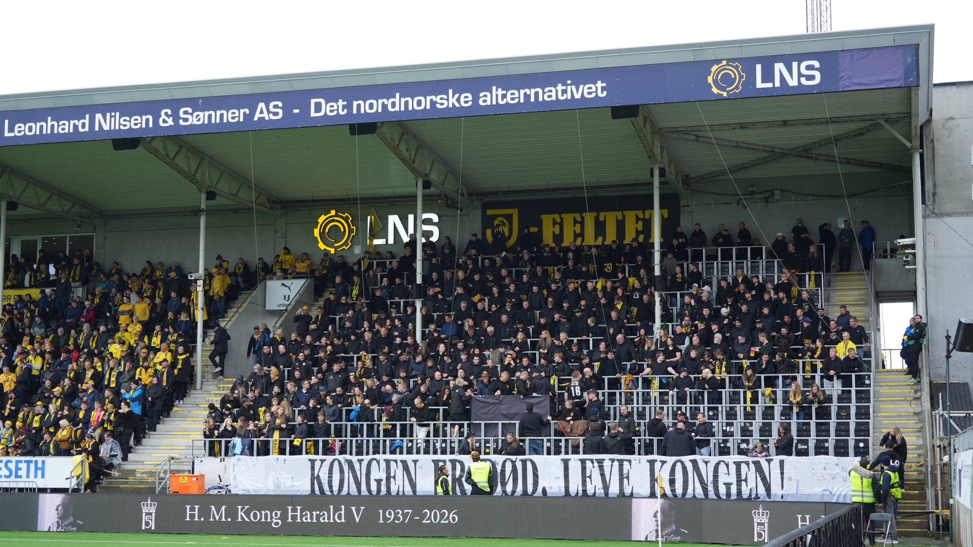 Supportere på en fotballtribune med et banner som hedrer Kong Harald V. (Bildebeskrivelsen er laget av en KI-tjeneste)