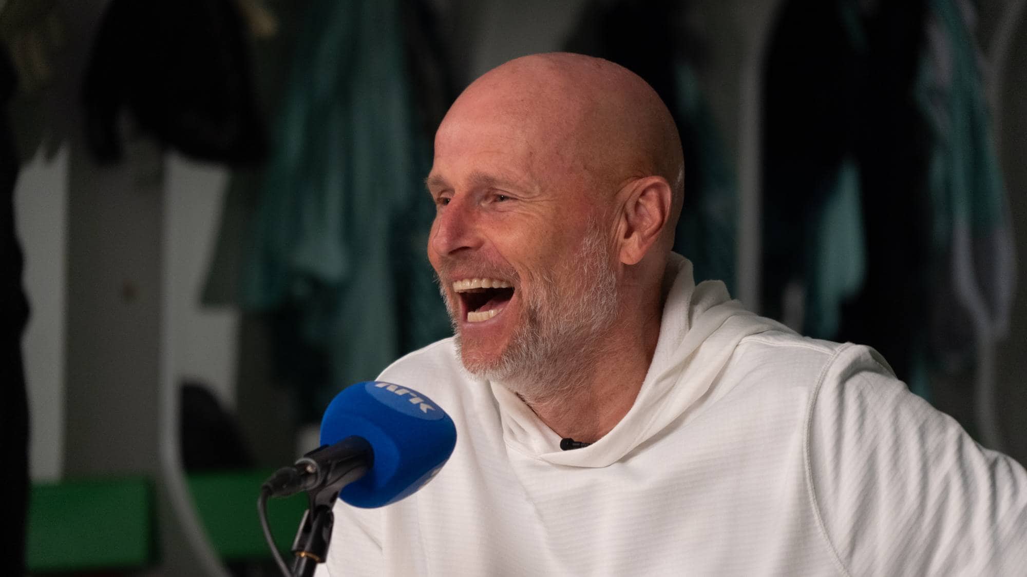 Ståle Solbakken ler mens han snakker i en mikrofon med NRK-logo i et garderobemiljø. (Bildebeskrivelsen er laget av en KI-tjeneste)