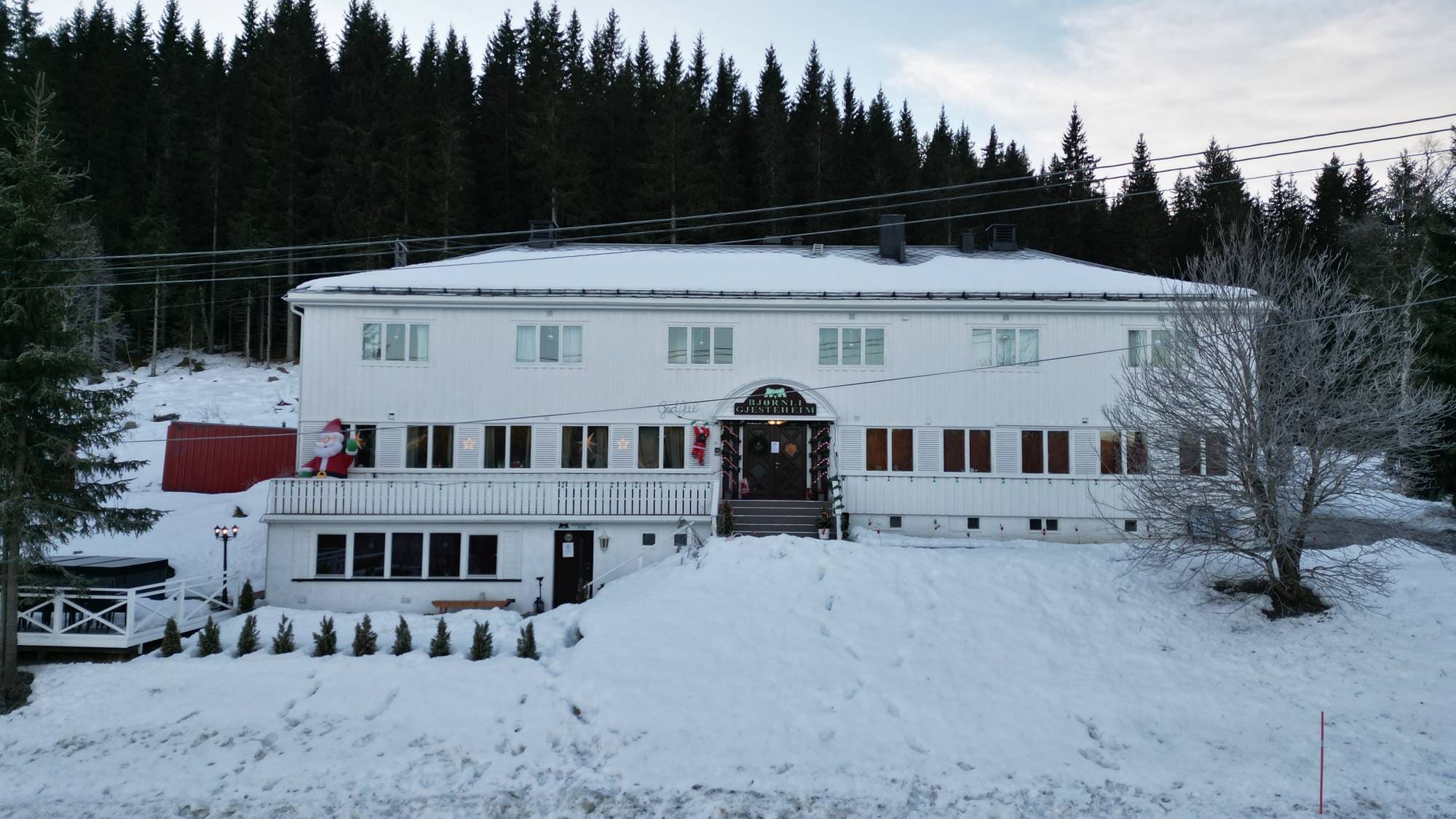 Bildet viser en hvit bygning beliggende i et snødekt landskap, med høye grantrær i bakgrunnen. Bygningen ser ut til å være en restaurant eller et overnattingssted, med flere vinduer og en stor inngangsdør. Utenfor er det dekorasjoner, inkludert julenisser, som tyder på høytid. Snøen dekker bakken, og det er spor etter tidligere ferdsel. (Bildebeskrivelsen er laget av en KI-tjeneste)