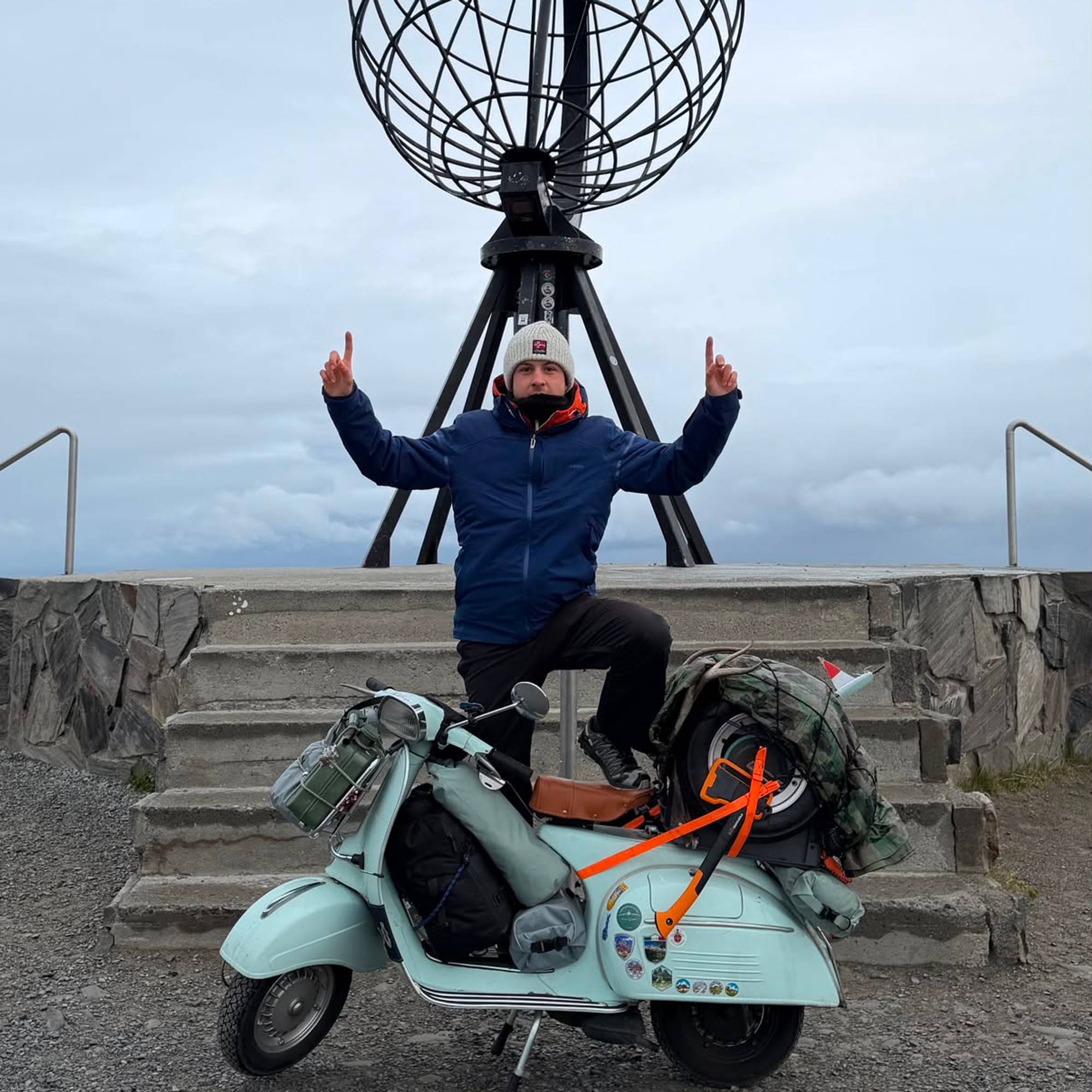 En mann står med et ben på en lys turkis vespa på Nordkapp