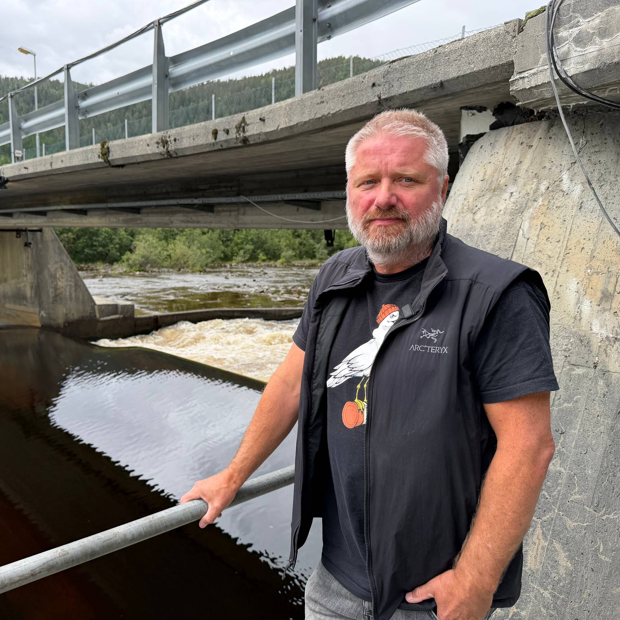 Mann med skjegg står ved et elveanlegg med en betongbro i bakgrunnen. (Bildebeskrivelsen er laget av en KI-tjeneste)