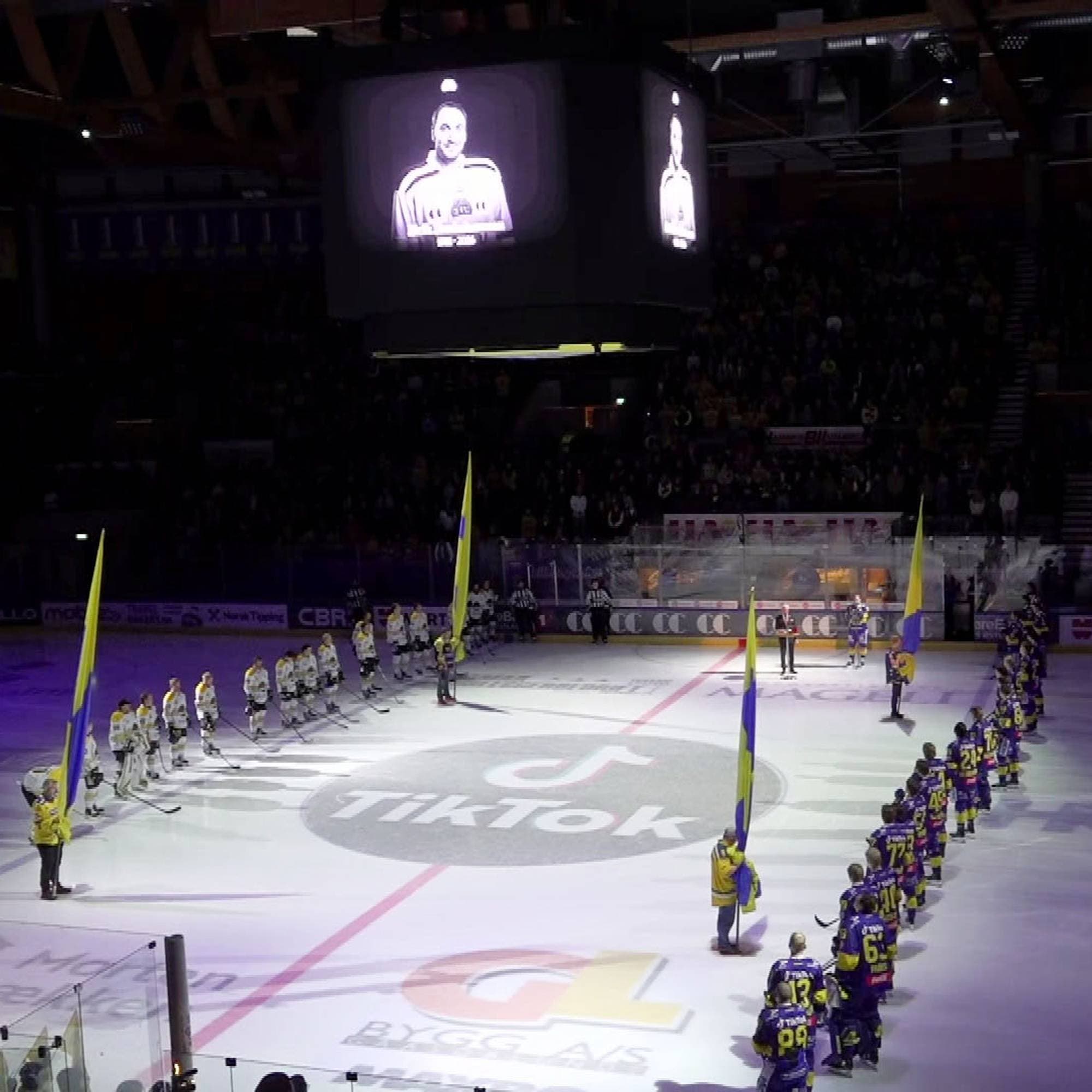Ishockeyspillere står oppstilt på isen i en arena før kampstart under et portrett på storskjermen. 