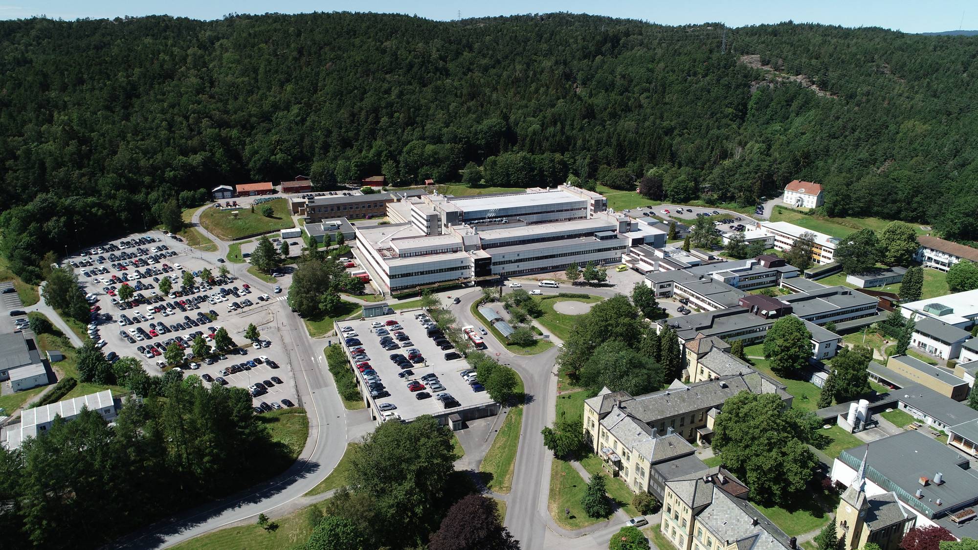 Dronebilde av et stort sykehuskompleks med flere bygninger. Det er mange biler parkert på et stort parkeringsområde til venstre i bildet. I bakgrunnen er det en tett skogkledd ås. Bygningene er moderne, med hvite og grå fasader, og ligger spredt i et grønt landskap. (Bildebeskrivelsen er laget av en KI-tjeneste)