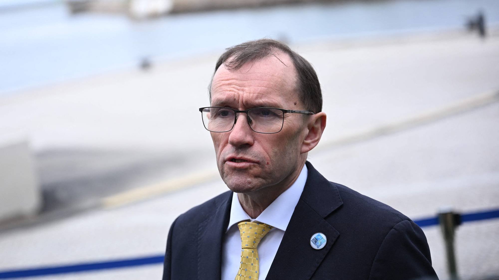 Utenriksminister Espen Barth Eide har på seg en mørk dressjakke, hvit skjorte og et gult slips.
På reversen av dressjakken sitter en knapp med et bilde på.
Bakgrunnen er ute ved sjøen med en kai. 
