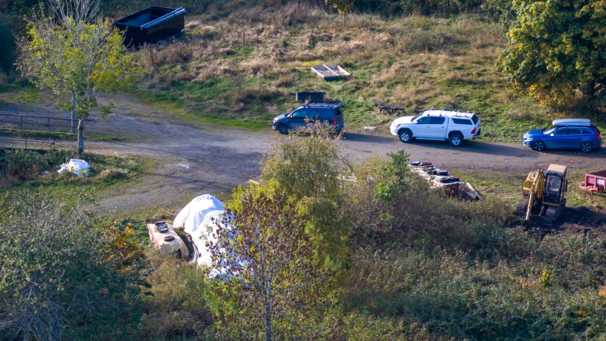 Flere biler og en gravemaskin står på et grusområde ved en åker og en rød låve. (Bildebeskrivelsen er laget av en KI-tjeneste)
