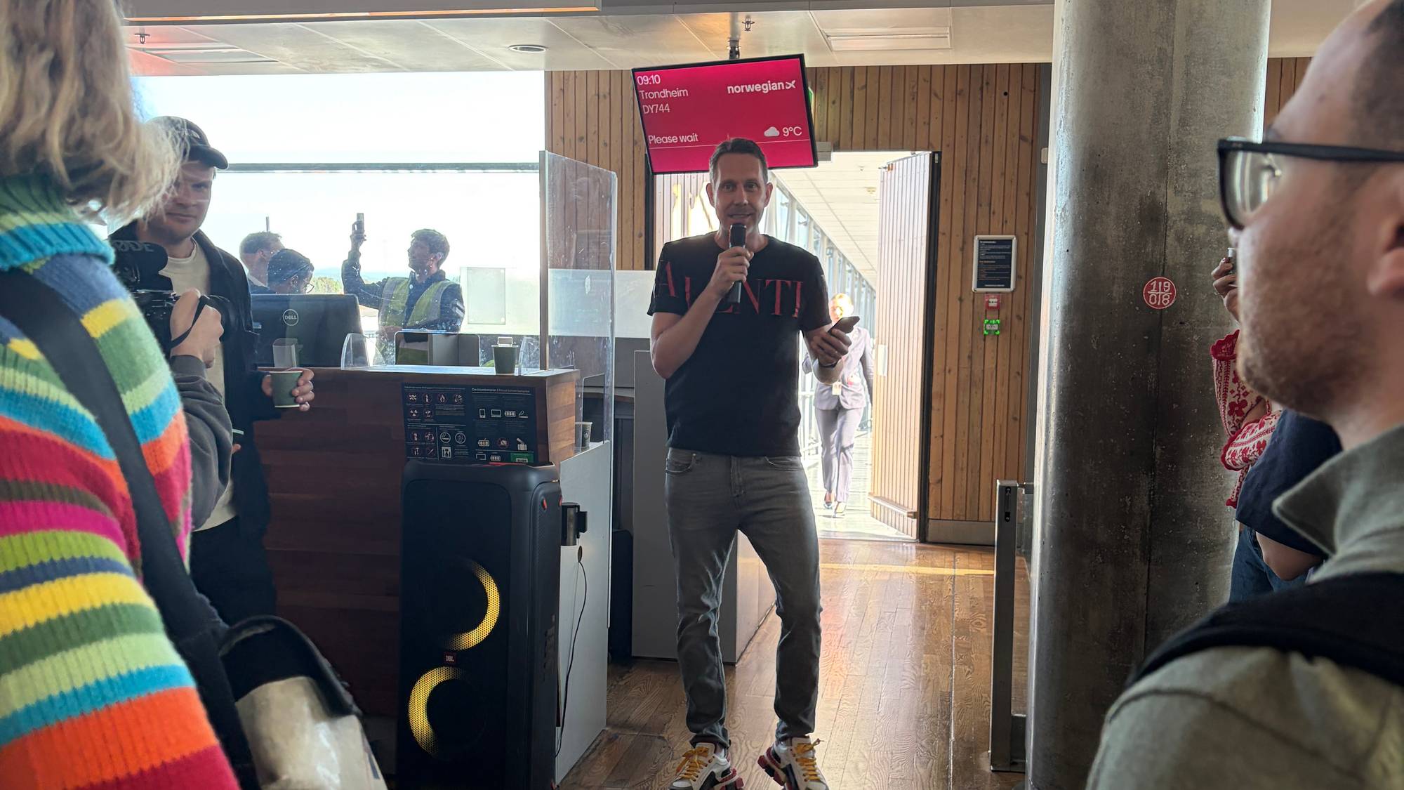 Komiker Morten Ramm står foran utgangen til en gate På Oslo lufthavn Gardermoen. Han her en mikrofon i hendene og ser glad ut. 