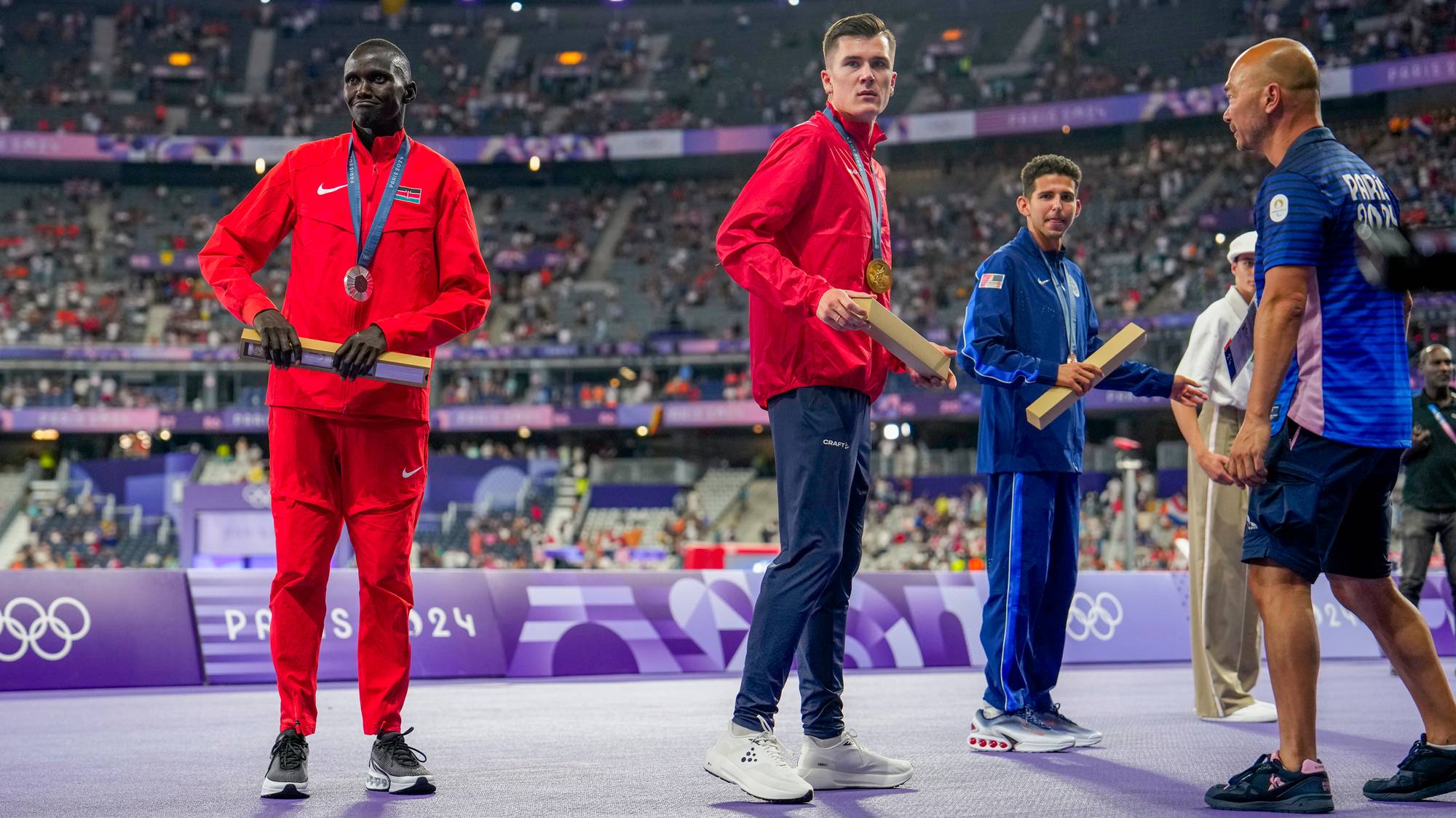 Tre utøvere står på et podium. Utøveren til venstre har på seg rød drakt og har medalje rundt halsen. Utøveren i midten har på seg rød jakke og gullmedalje. Utøveren til høyre er iført blå drakt. Publikum sitter på tribunene bak dem. (Bildebeskrivelsen er laget av en KI-tjeneste)