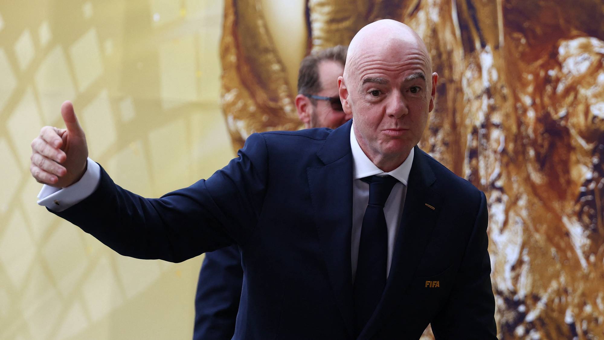 FIFA-president Gianni Infantino i mørk dress med utstrakt hånd foran en gullfarget bakgrunn. (Bildebeskrivelsen er laget av en KI-tjeneste)