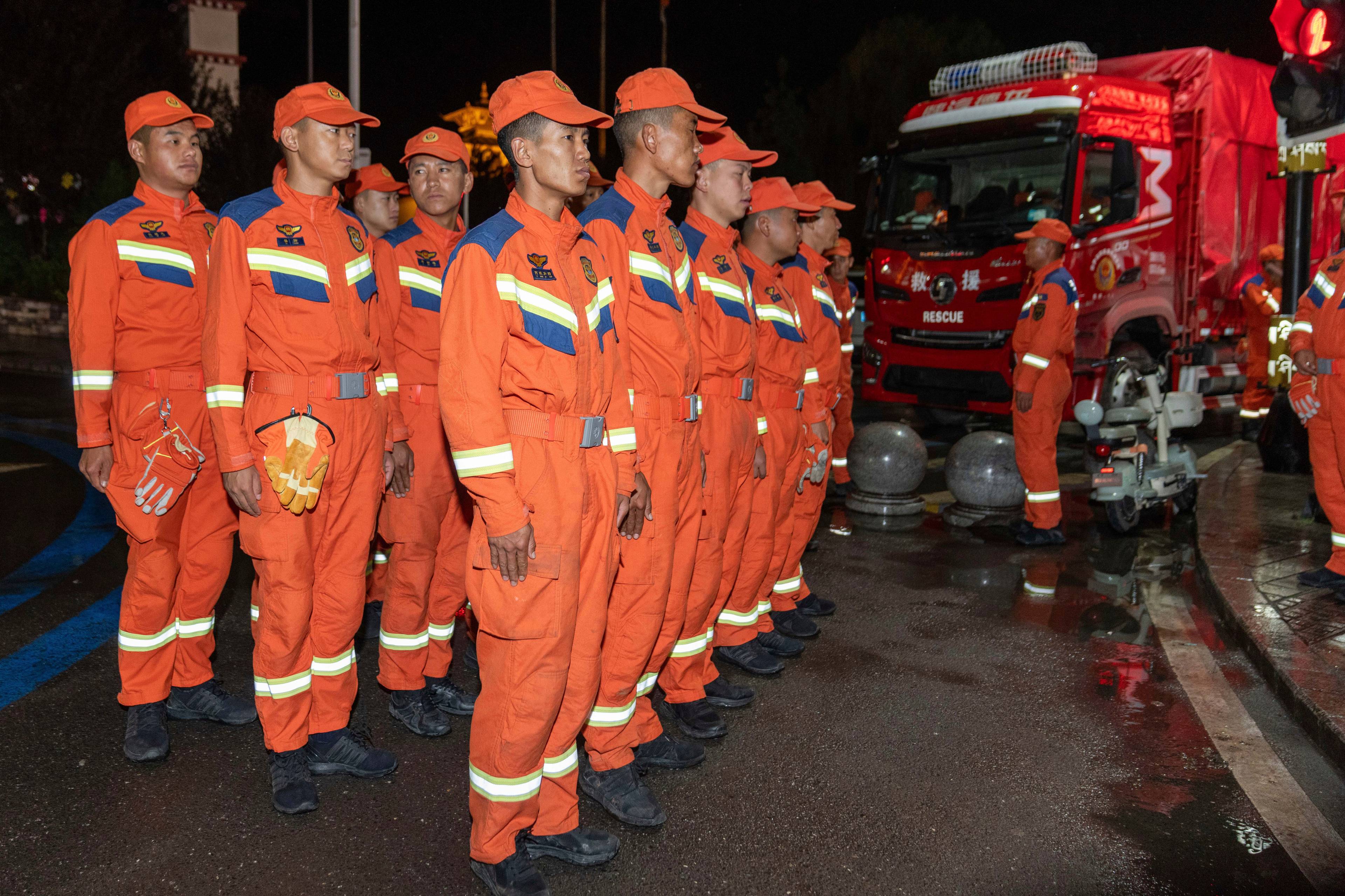 Redningsarbeidere i oransje uniformer står oppstilt ved en rød brannbil om kvelden. (Bildebeskrivelsen er laget av en KI-tjeneste)
