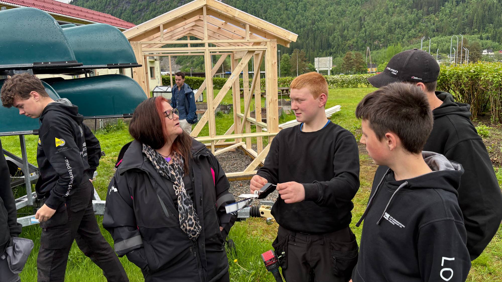 En gruppe ungdommer og en voksen kvinne ser på et trebygg som er under oppføring. Bak dem er det en rekke med kanoer på en henger. Landskapet er preget av grønne fjellsider med tåke. (Bildebeskrivelsen er laget av en KI-tjeneste)