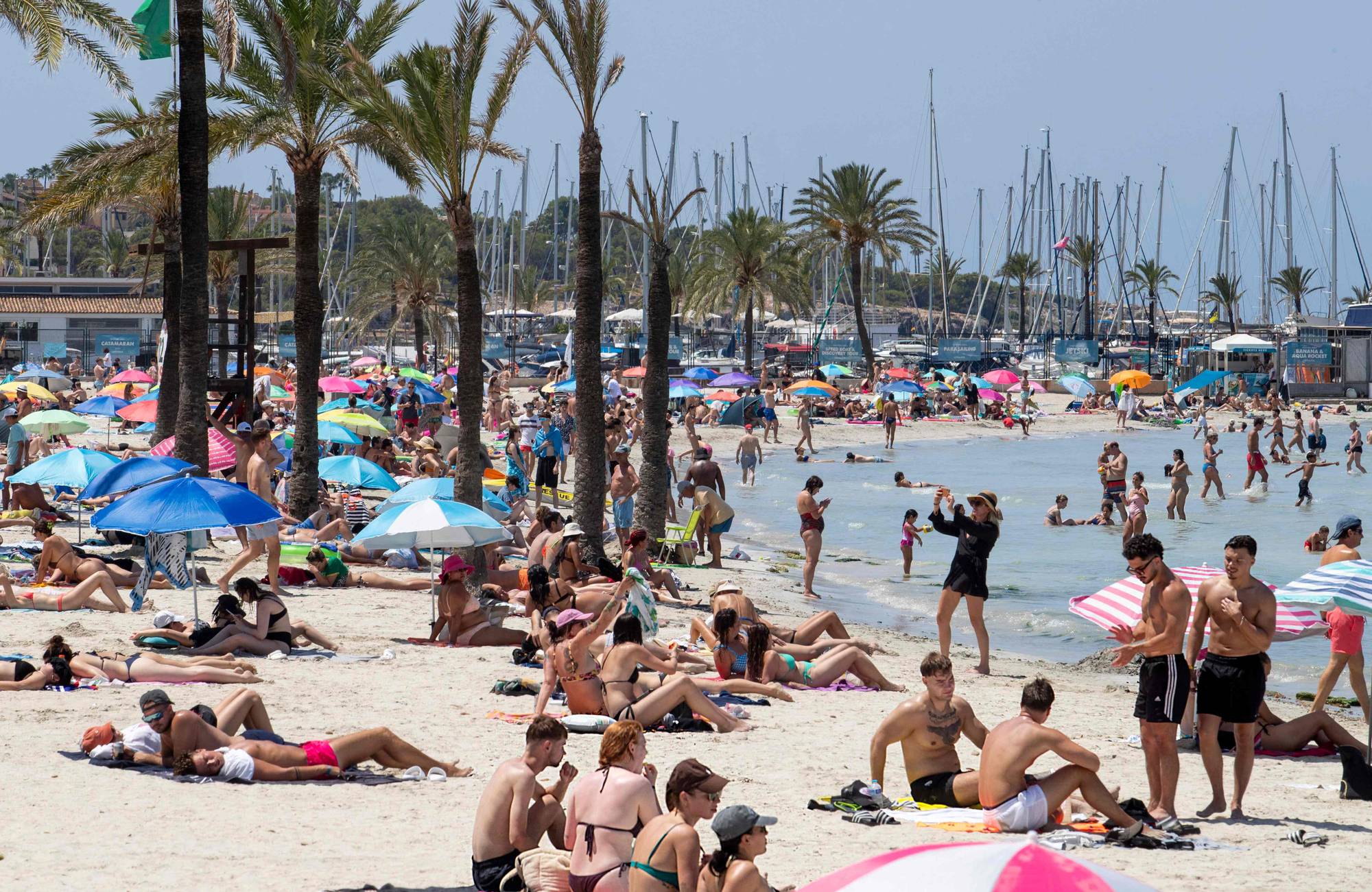 Strand med mange turister som soler seg og bader. Hvit sand, vann og palmer. 
