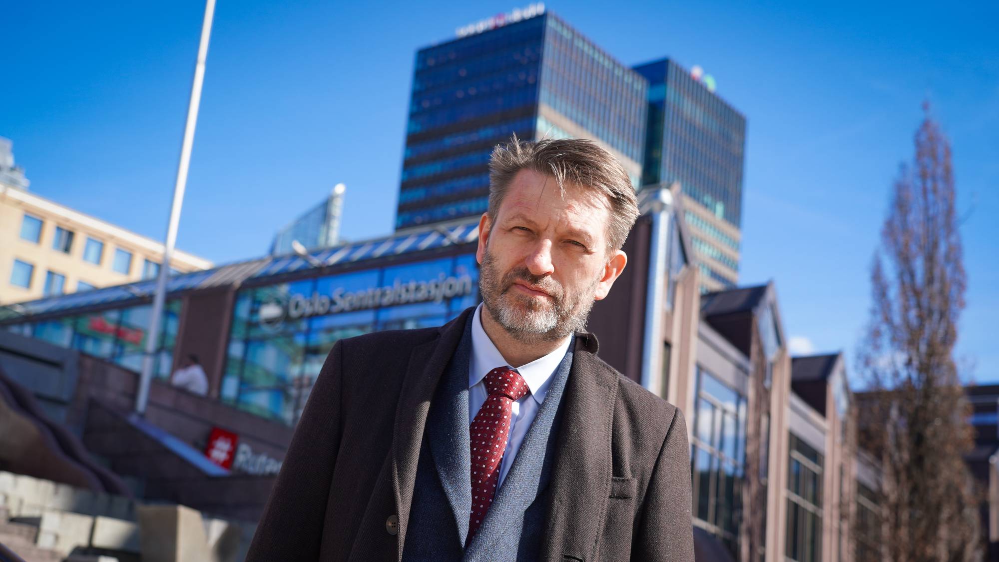 En mann med grått skjegg står foran Oslo S. Han har på seg mørk frakk, vest, hvit skjorte og et rødt slips med hvite prikker. Bak mannen ses flere bygninger, deriblant et glassbygg med teksten "Oslo Sentralstasjon". (Bildebeskrivelsen er laget av en KI-tjeneste)