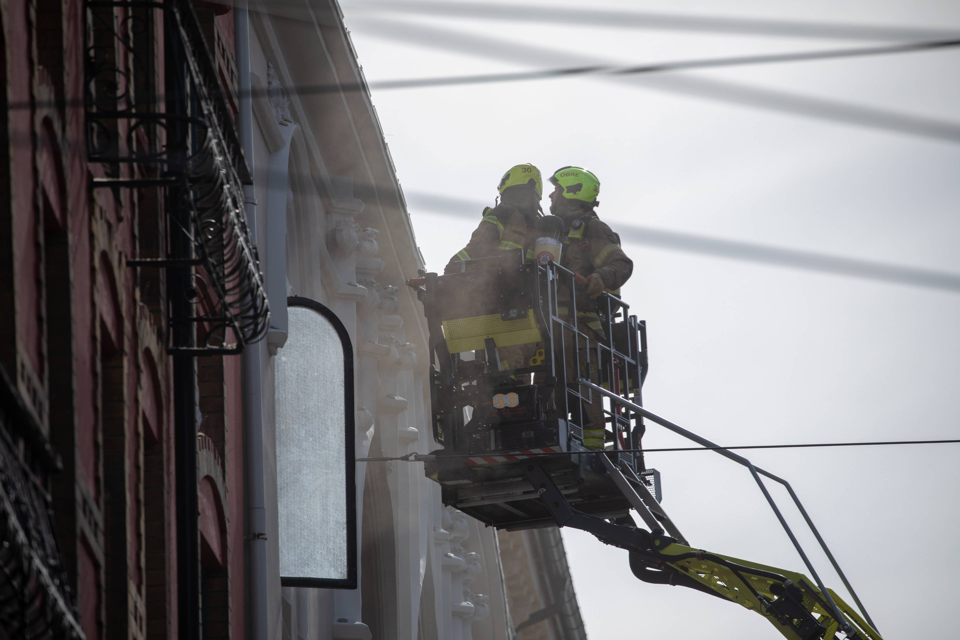 To brannmenn i lift undersøker en bygningsfasade. De er iført verneutstyr, inkludert gule hjelmer. Mannskapet befinner seg på et byggegjerde. (Bildebeskrivelsen er laget av en KI-tjeneste)