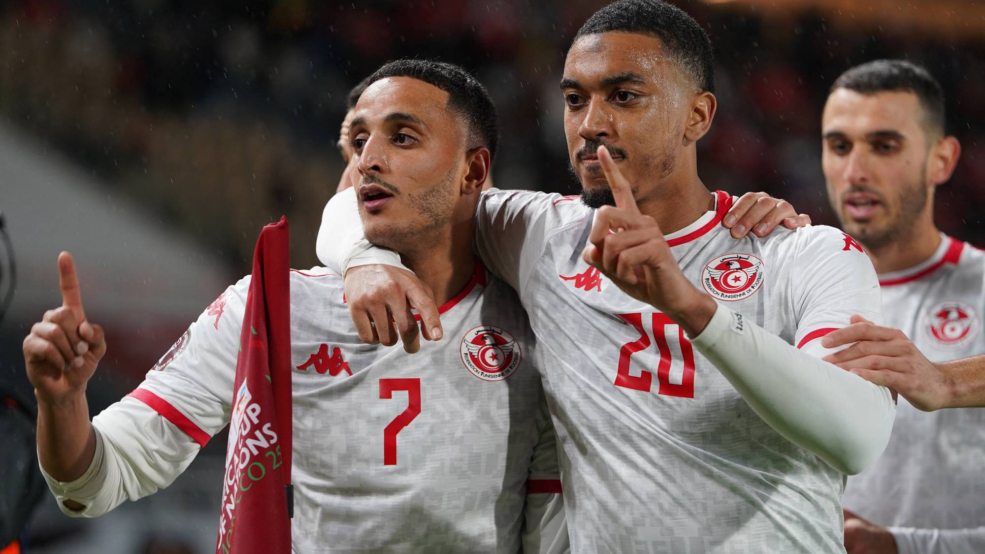To fotballspillere feirer sammen under en kamp. Spilleren til venstre, med trøye nummer 7, har en arm rundt skulderen til spilleren til høyre med trøye nummer 20. Begge peker opp med en finger. En annen spiller er synlig i bakgrunnen til høyre. Spillerne er ikledd hvite landslagsdrakter. (Bildebeskrivelsen er laget av en KI-tjeneste)