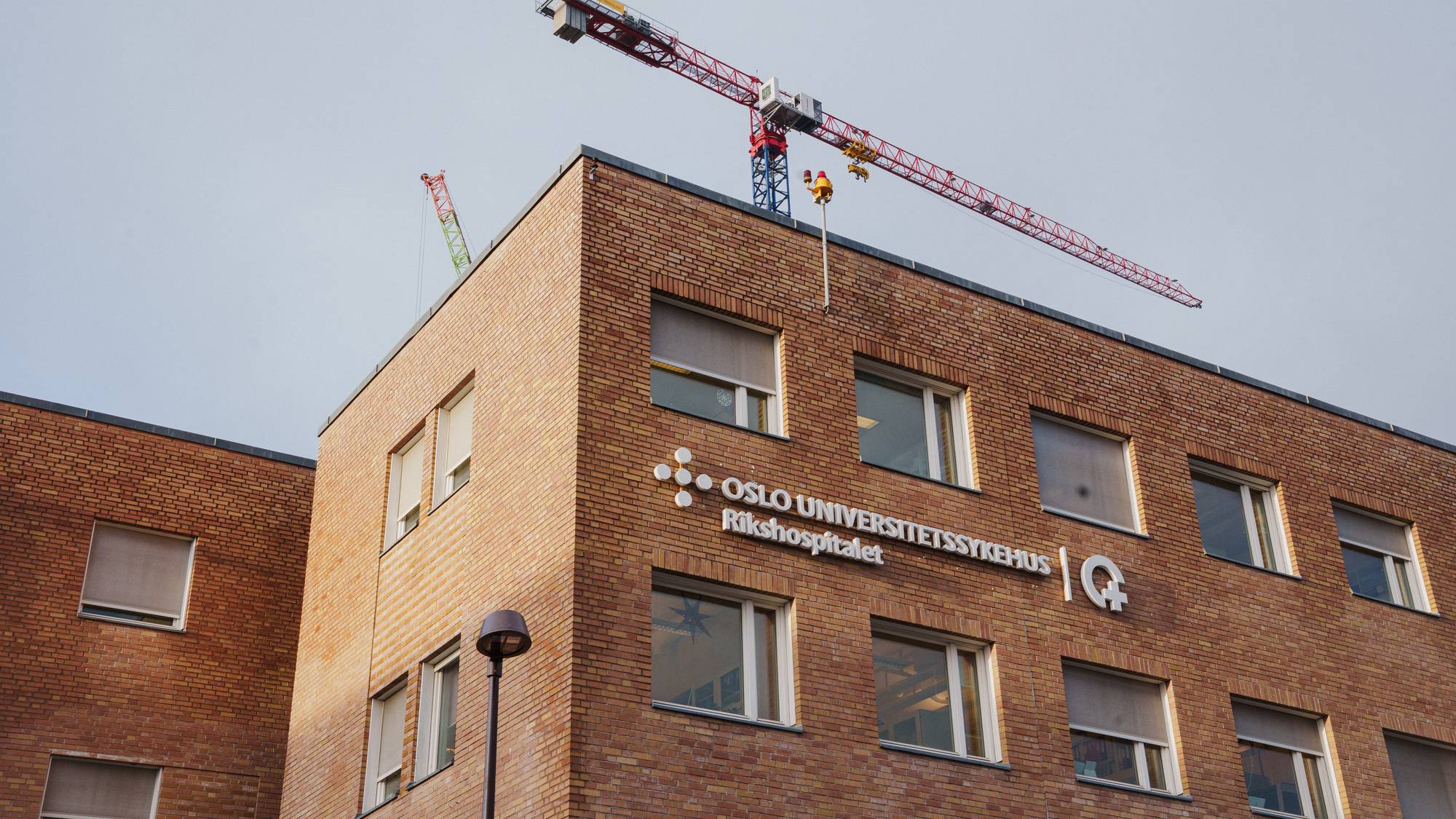 En stor byggekran strekker seg over taket på Oslo universitetssykehus, Rikshospitalet. Bygningen er kledd i murstein, og det er flere vinduer på fasaden. En mindre kran er synlig i bakgrunnen. Været er skyet. (Bildebeskrivelsen er laget av en KI-tjeneste)