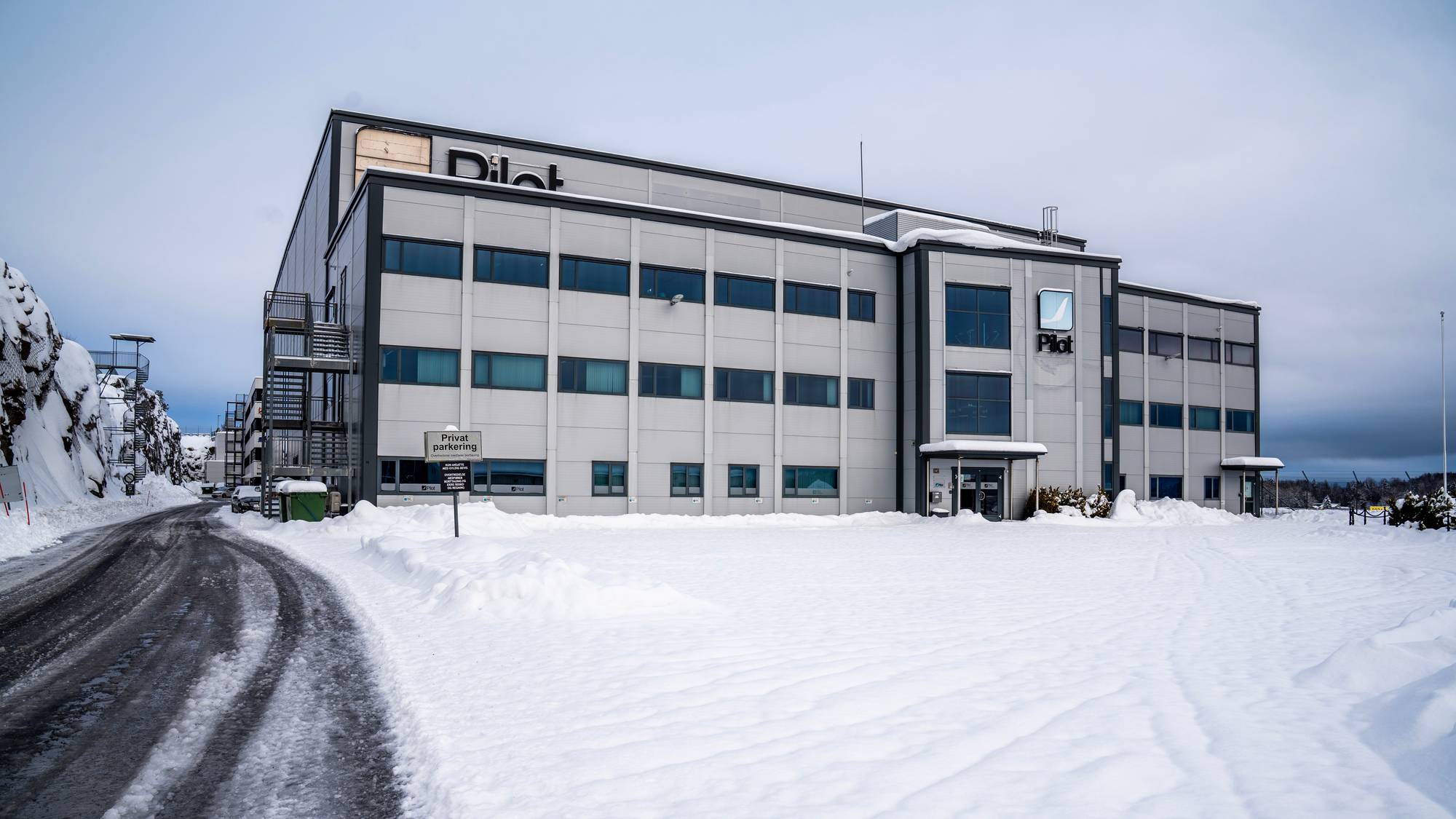 Bildet viser en stor, modern bygning med navnet "Pilot" i tydelig skrift på fasaden. Bygningen står på en snødekt tomt, og det er spor etter kjøretøy i snøen som omkranser en asfaltvei. Omgivelsene inkluderer bratte snøfylte skråninger på begge sider av veien. Det henger skilt ved inngangen som angir privat parkering.