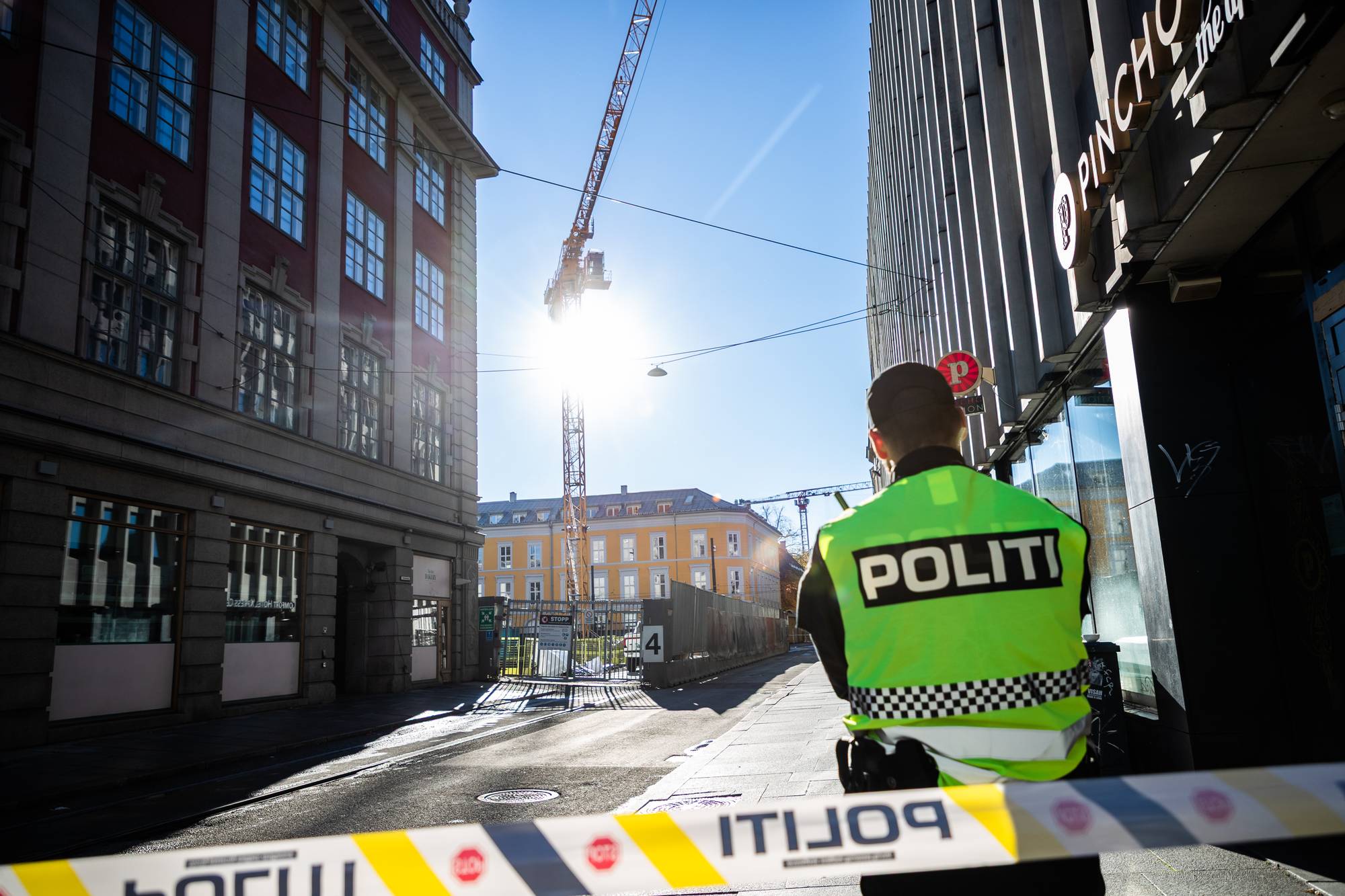 Politibetjent med refleksvest står ved sperrebånd i en bygate. Bygårder og byggekran i bakgrunnen. (Bildebeskrivelsen er laget av en KI-tjeneste)