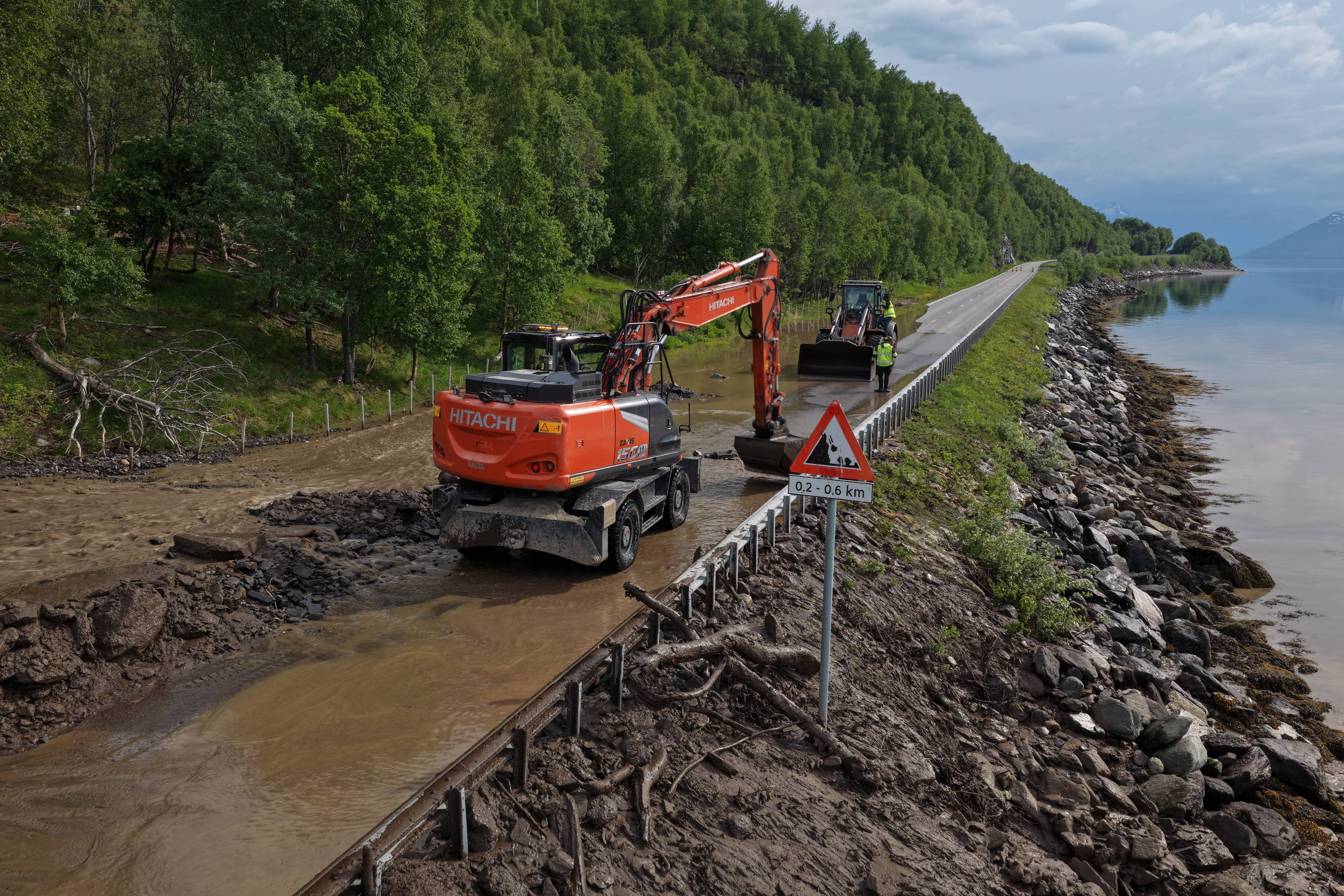 Gravmaskiner rydder veien etter flomskader. To maskinførere i gule vester overvåker arbeidet. Et fareskilt for rasvarsel står ved veikanten. Veien går langs en innsjø med fjell i bakgrunnen. (Bildebeskrivelsen er laget av en KI-tjeneste)