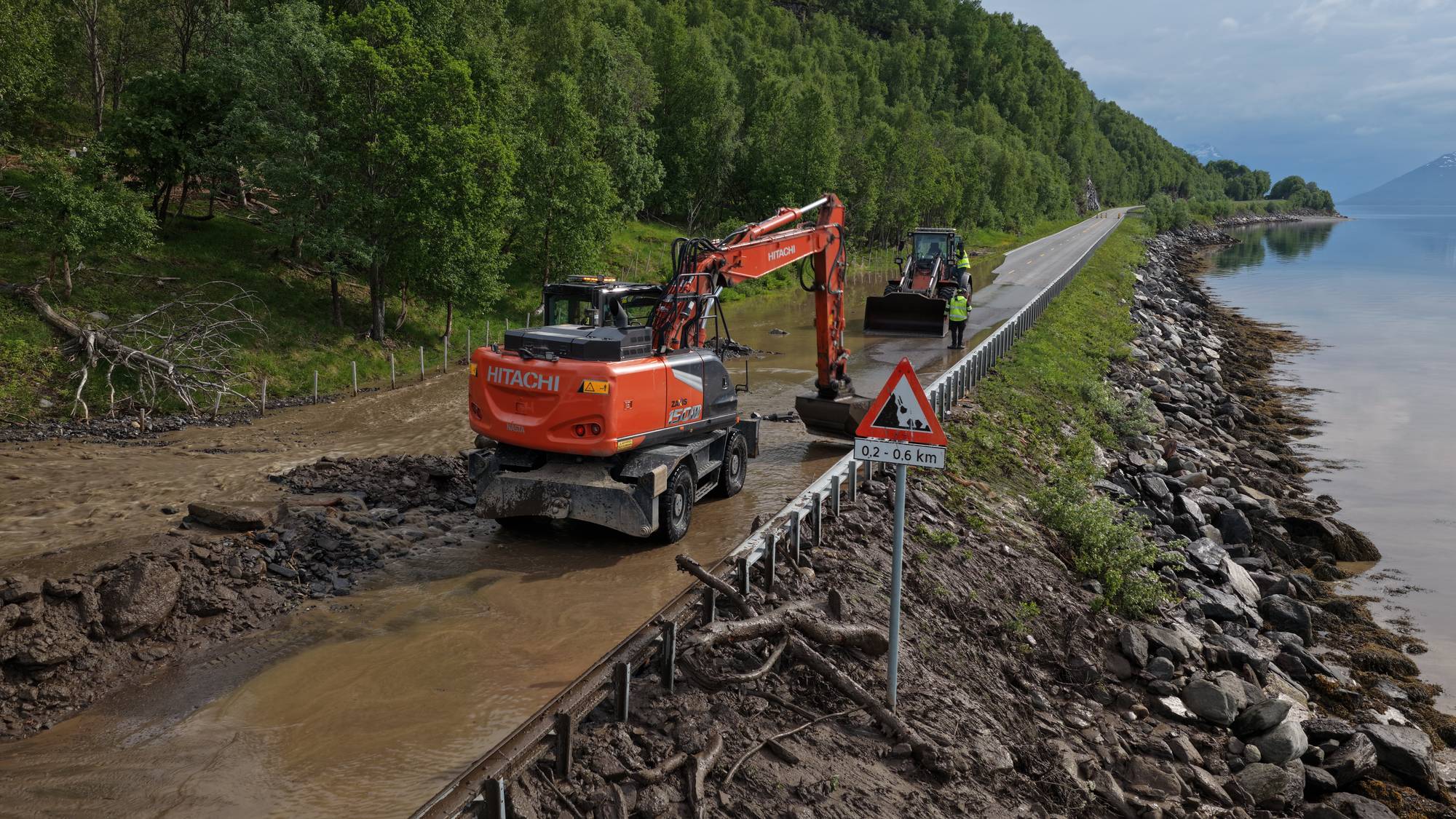 Gravmaskiner rydder veien etter flomskader. To maskinførere i gule vester overvåker arbeidet. Et fareskilt for rasvarsel står ved veikanten. Veien går langs en innsjø med fjell i bakgrunnen. (Bildebeskrivelsen er laget av en KI-tjeneste)