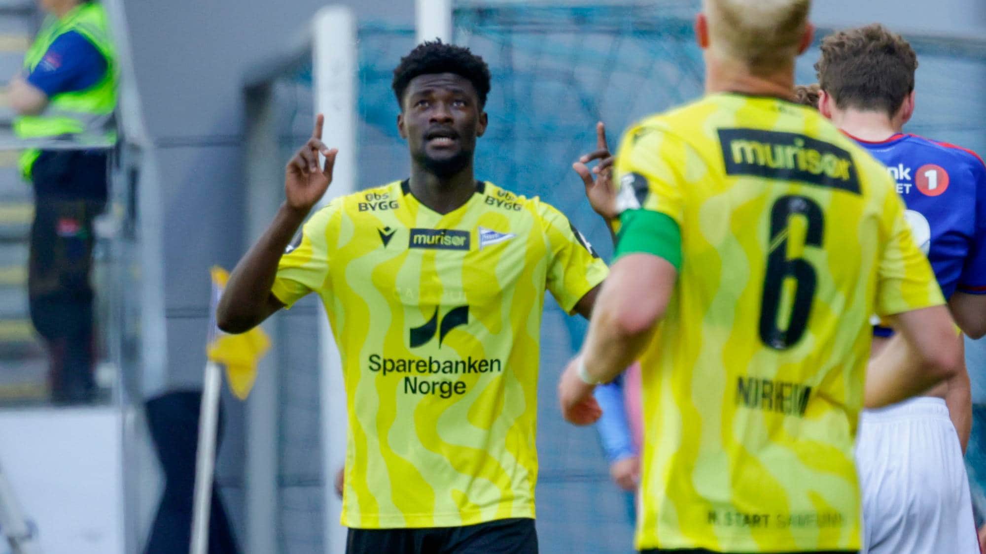 Fotballspiller med nummer 10 på drakten hever hendene. Han har på seg gul trøye med grønn krage. En annen spiller med nummer 6 på ryggen står foran ham. Det er en fotballkamp. (Bildebeskrivelsen er laget av en KI-tjeneste)