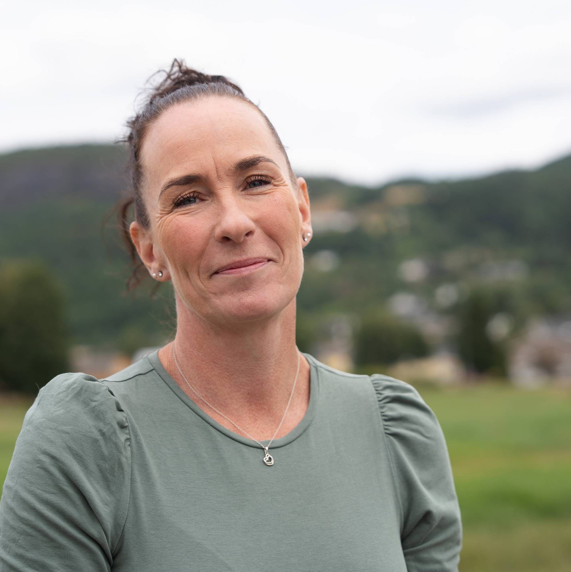 Kvinne med oppsatt hår i grønn topp utendørs med landskap i bakgrunnen. (Bildebeskrivelsen er laget av en KI-tjeneste)