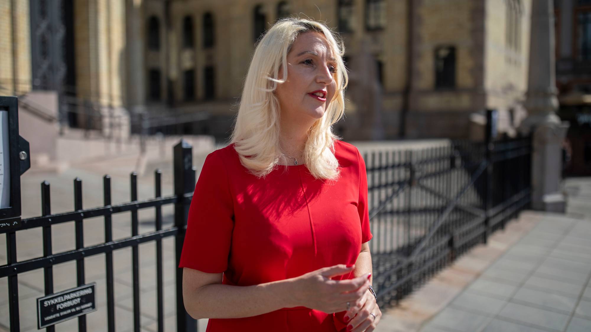 Kvinne med lyst hår i rød kjole står utendørs foran Stortinget. 