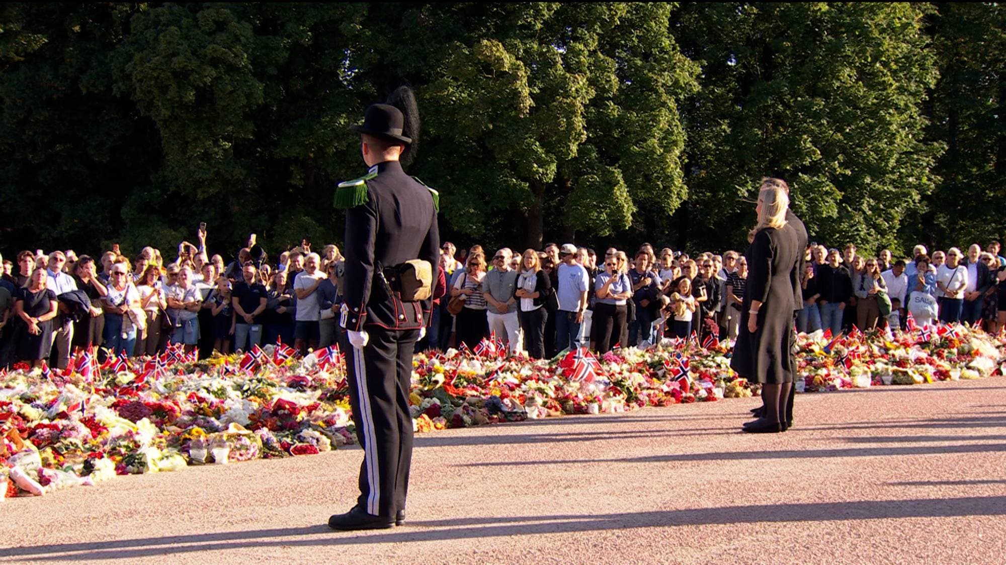 Kongefamilien ved blomsterhavet utenfor Slottet med en gardist og folkemengde. (Bildebeskrivelsen er laget av en KI-tjeneste)