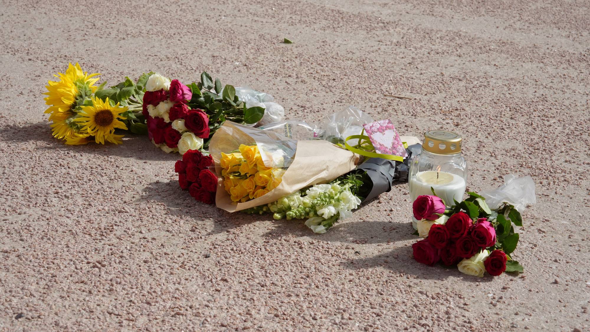 Blomsterbuketter og et tent lys plassert på et grusunderlag. (Bildebeskrivelsen er laget av en KI-tjeneste)
