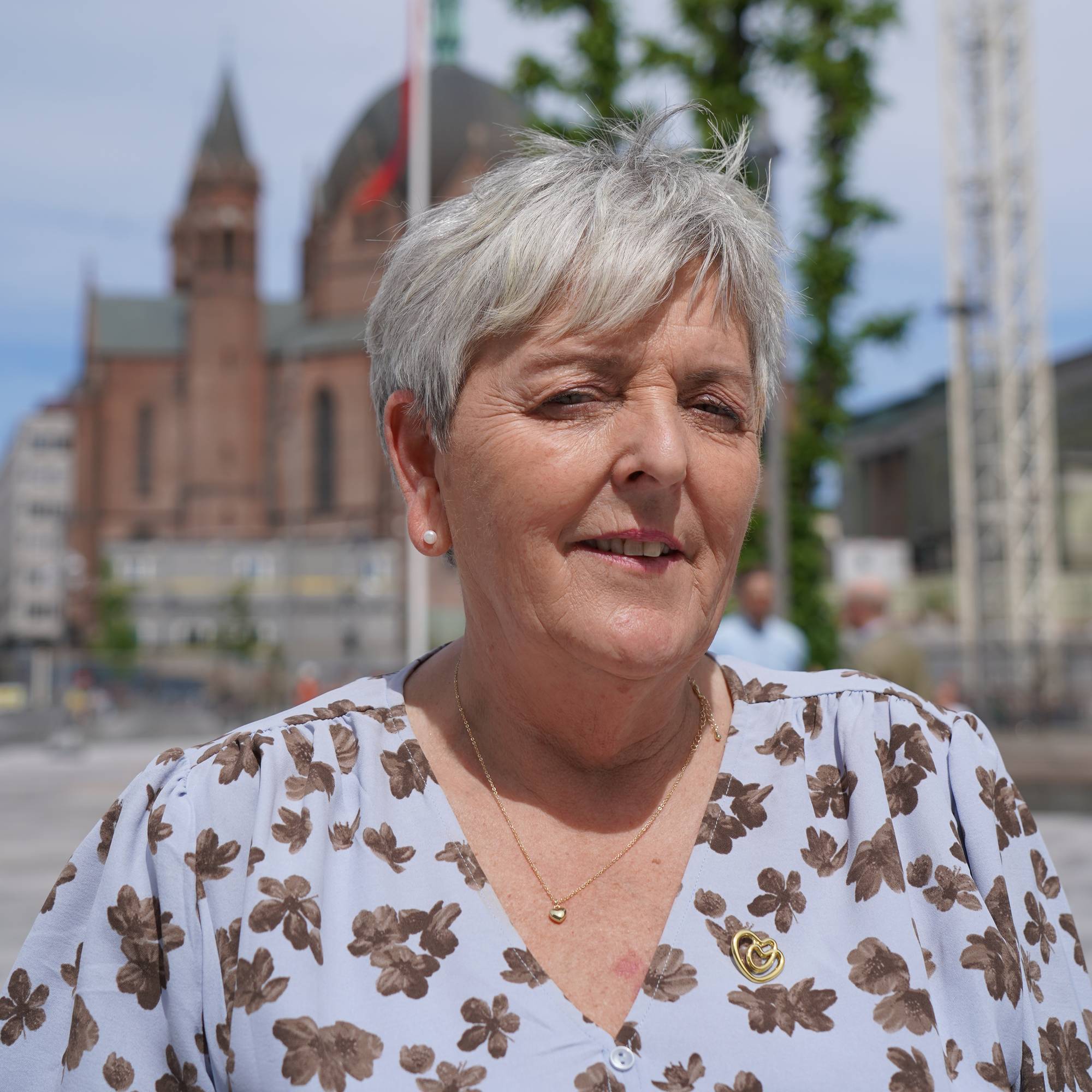 En eldre kvinne med grått hår og blomstermønstret bluse. Hun smiler og har et gullkjede med et hjerteanheng. I bakgrunnen ses en kirke og bygningsmasse under en klar himmel. Det er også en byggekran synlig. (Bildebeskrivelsen er laget av en KI-tjeneste)