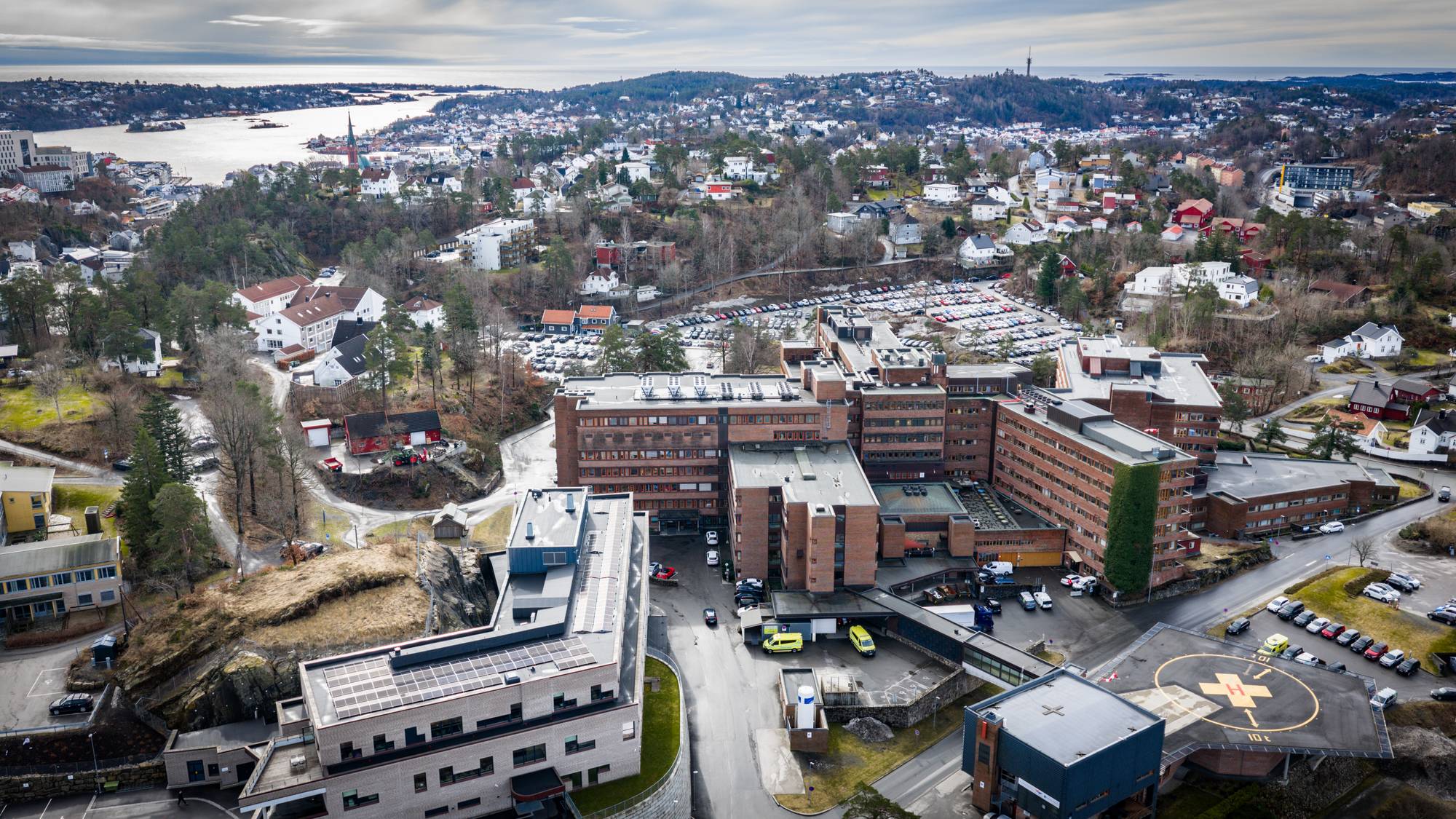 Et stort sykehusbygg i murstein sees fra luften. Det er en helikopterplass med en H foran sykehuset. Det er en stor parkeringsplass fylt med biler. Omgivelsene består av tettbebyggelse, trær og en vik. (Bildebeskrivelsen er laget av en KI-tjeneste)