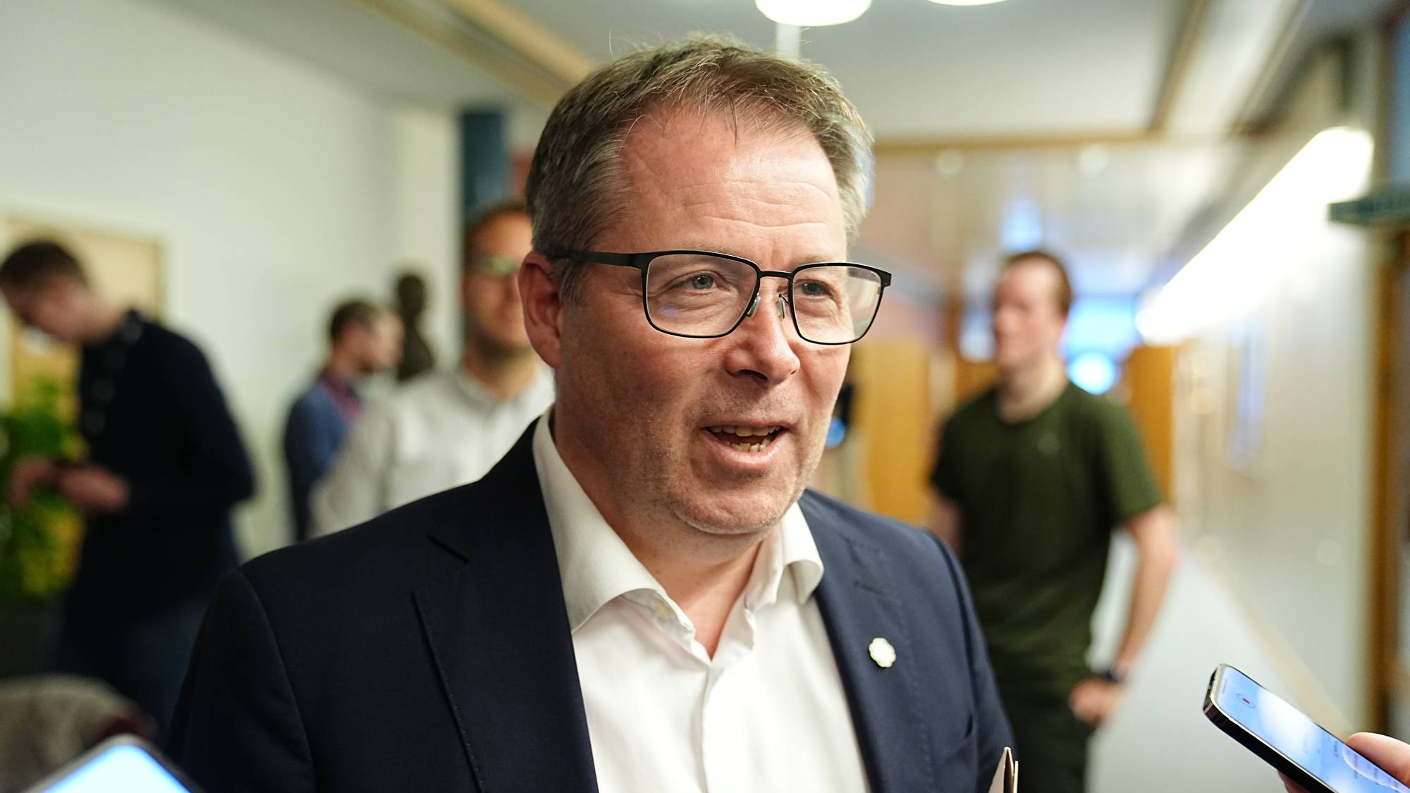 Bjørn Arild Gram i dress gir en uttalelse til flere journalister som holder mobiltelefoner. I bakgrunnen står flere personer og ser på. Omgivelsene er en bred korridor med moderne interiør. Det er god belysning og plantemateriale i rommet.