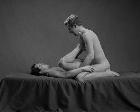 To nakne menn ligger over hverandre på en grå seng. Den nederste med føttene på den andres bryst