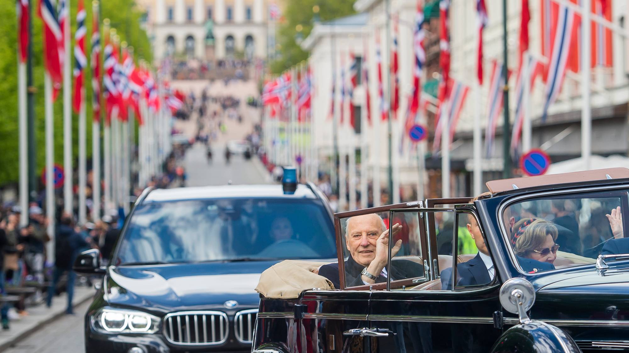 Kong Harald og dronning Sonja sitter i åpen bil og vinker i bilen A1 som kjøres nedover Karl Johan på 17. mai 2020