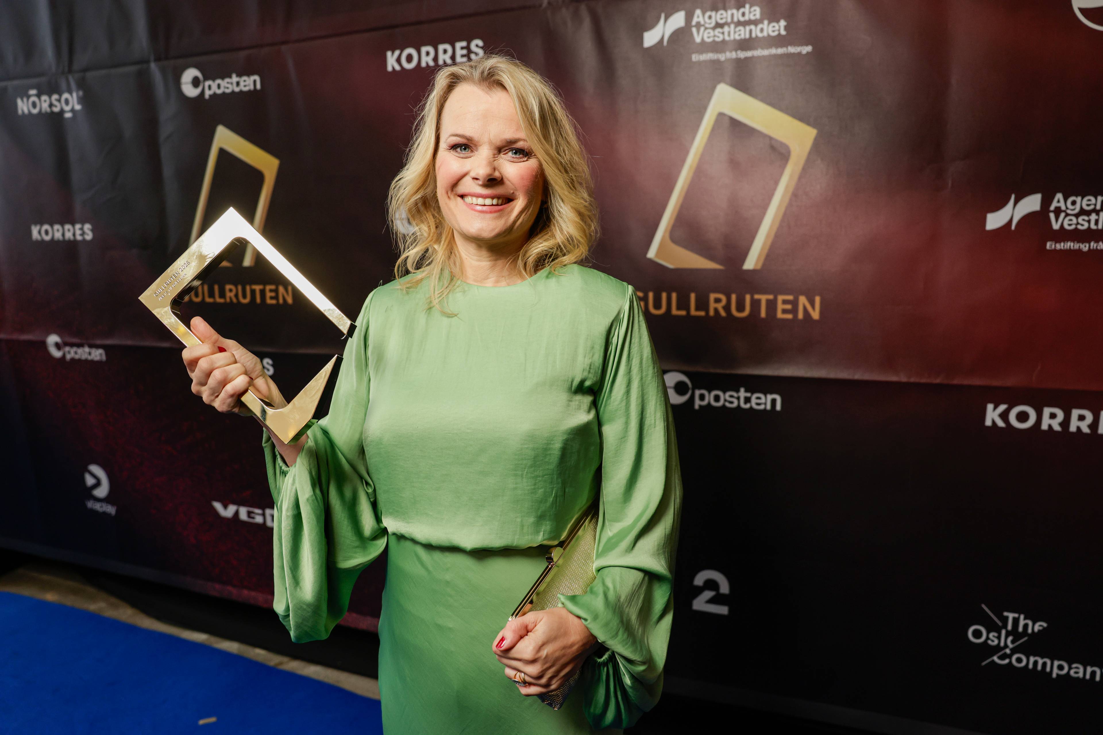 En kvinne i mintgrønn kjole holder en gullpris under en prisutdeling. Bak henne er en sponsorvegg med flere logoer. Kvinnen smiler til kamera. (Bildebeskrivelsen er laget av en KI-tjeneste)