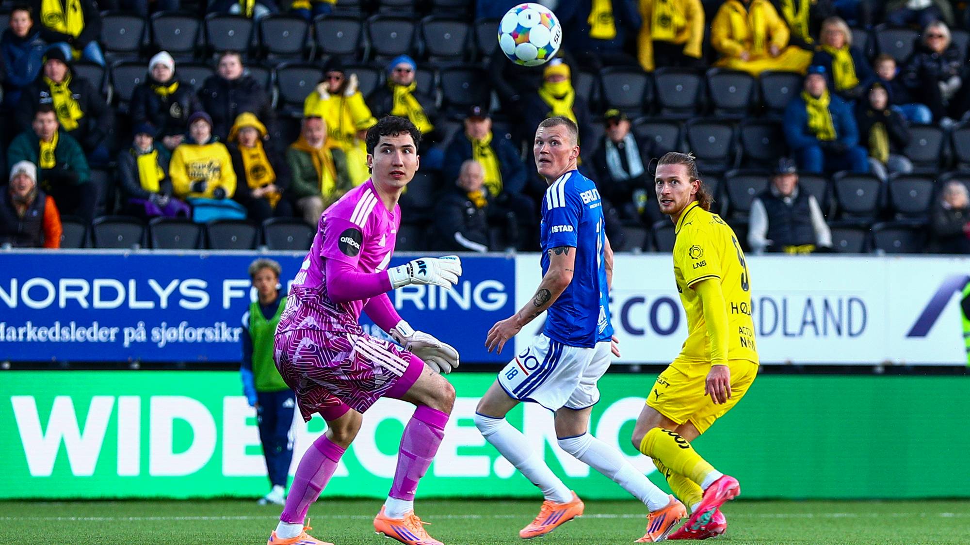Fotballspillere i aksjon under en kamp. En keeper i lilla drakt og to utespillere. Tilskuere sitter på tribunen i bakgrunnen. (Bildebeskrivelsen er laget av en KI-tjeneste)